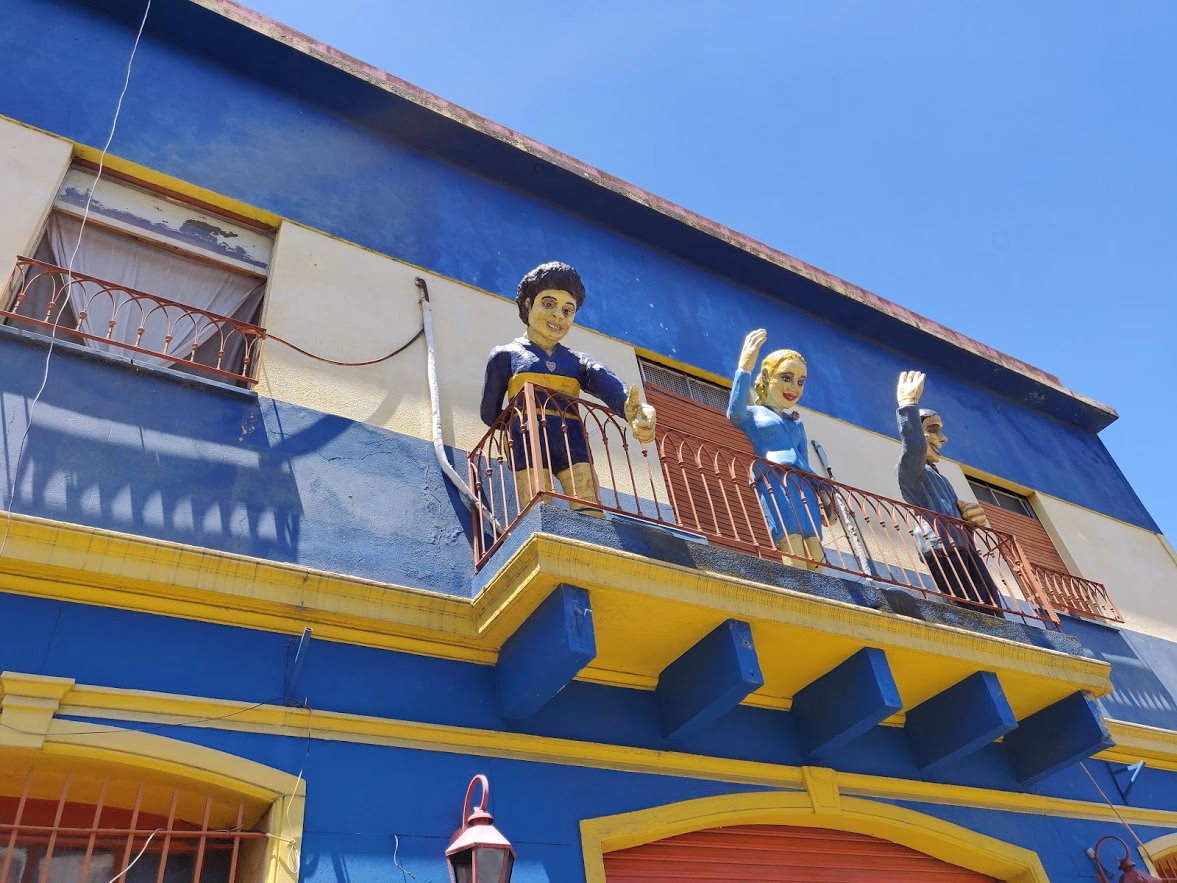 Life-sized statues of Maradona in a Boca jersey, Carlos Gardel in a fedora and Eva Perón wave from a blue-and-yellow balcony — the club colours painted straight onto the wall.