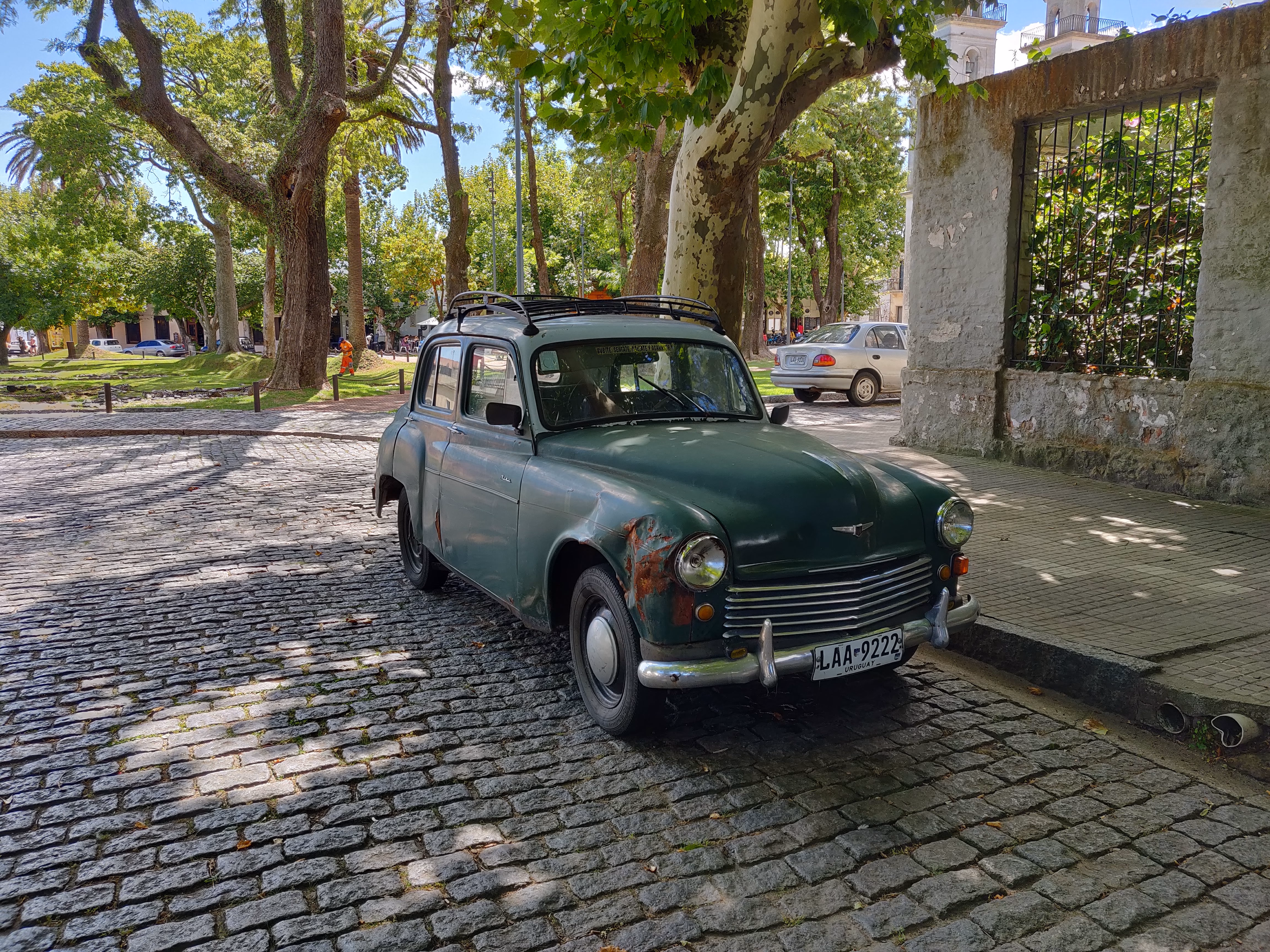A forest-green sedan with a roof rack beside a stone wall — wrought-iron window grilles and dappled light on the cobblestones.
