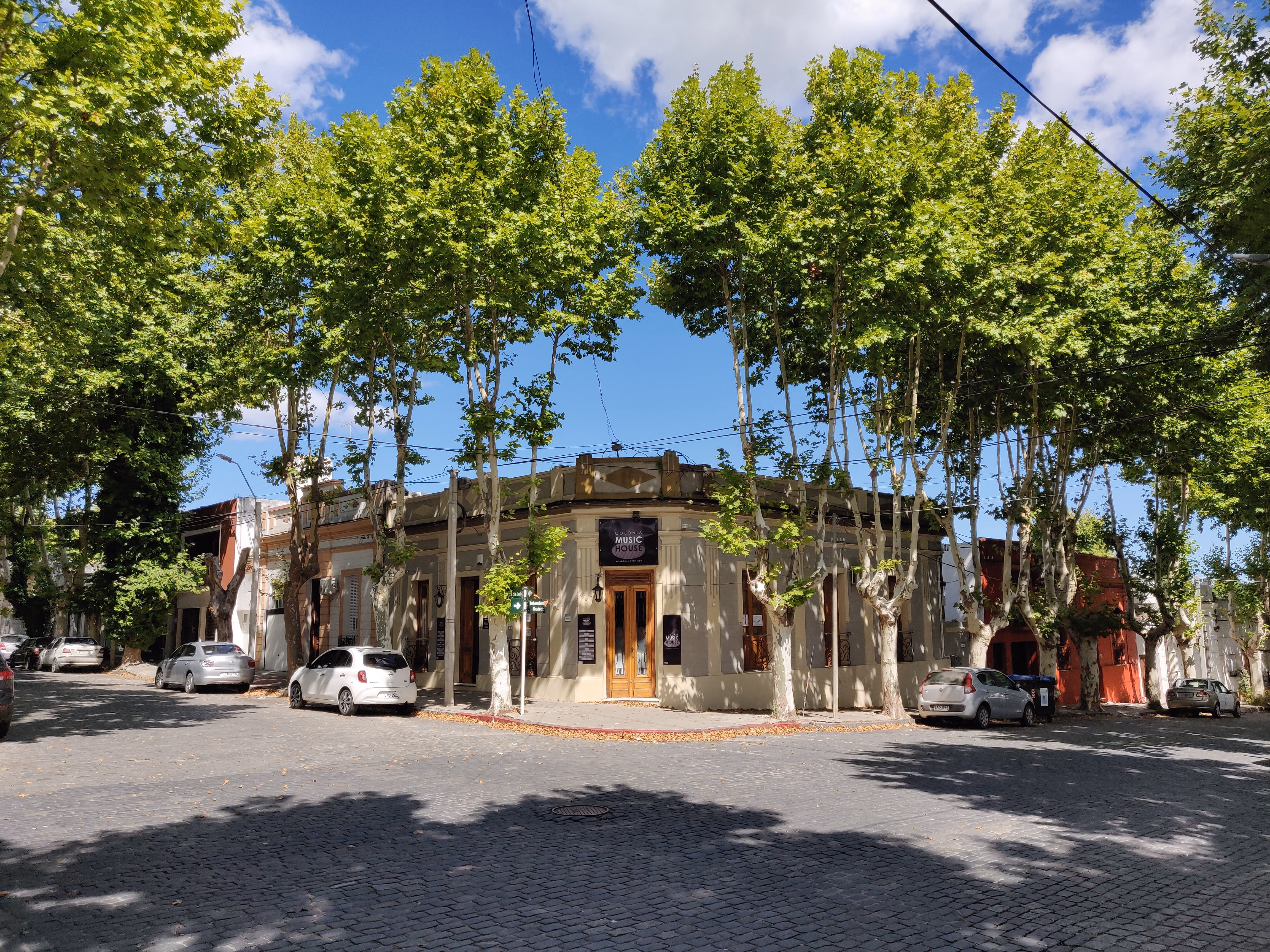 A rounded corner building under plane trees — "Colonia Music House" above a wooden door, cobbles fanning out across the intersection.
