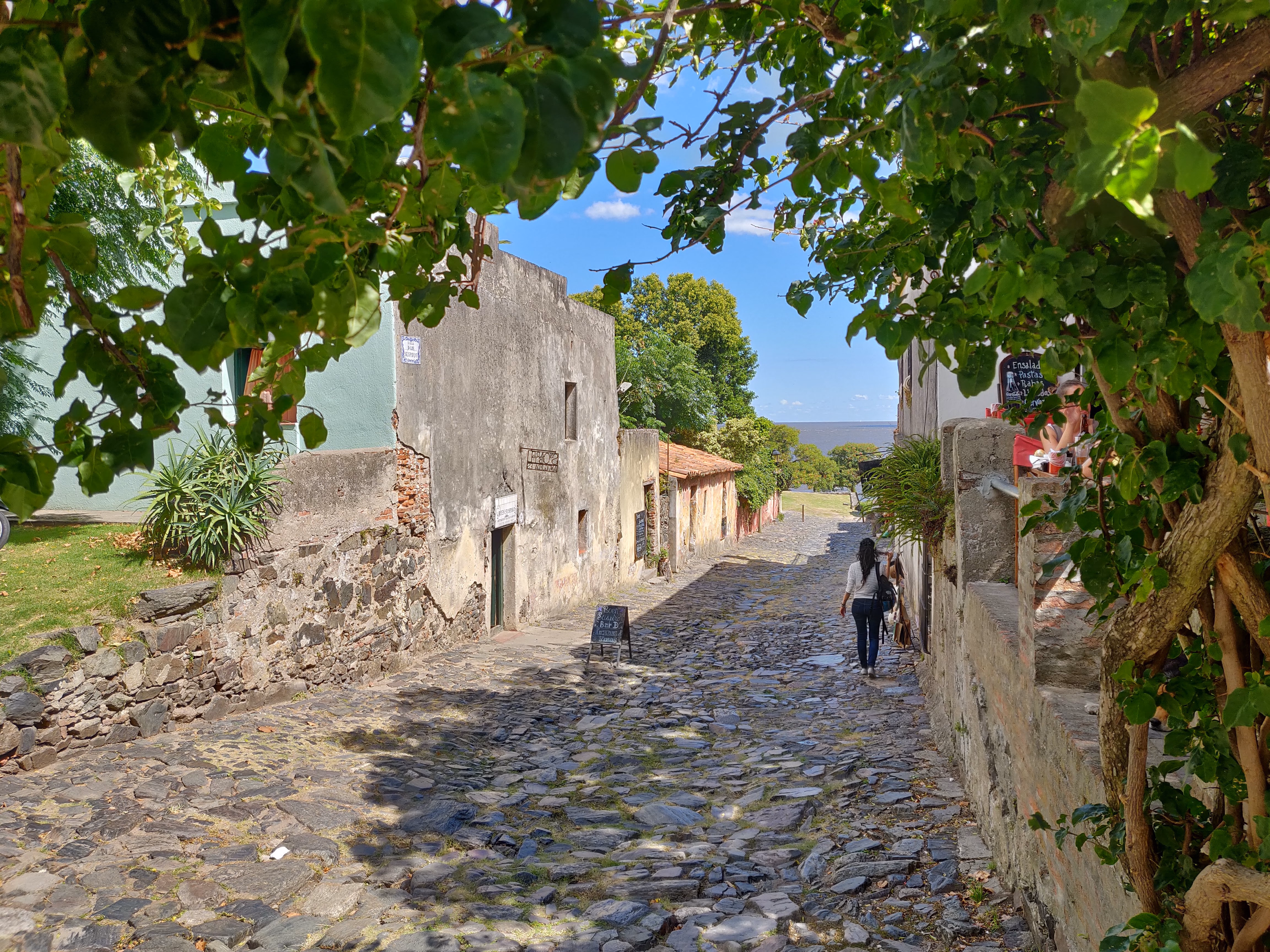 A cobbled street sloping toward the Río de la Plata — pastel green stucco, stone walls and café chairs under the trees at the water's edge.