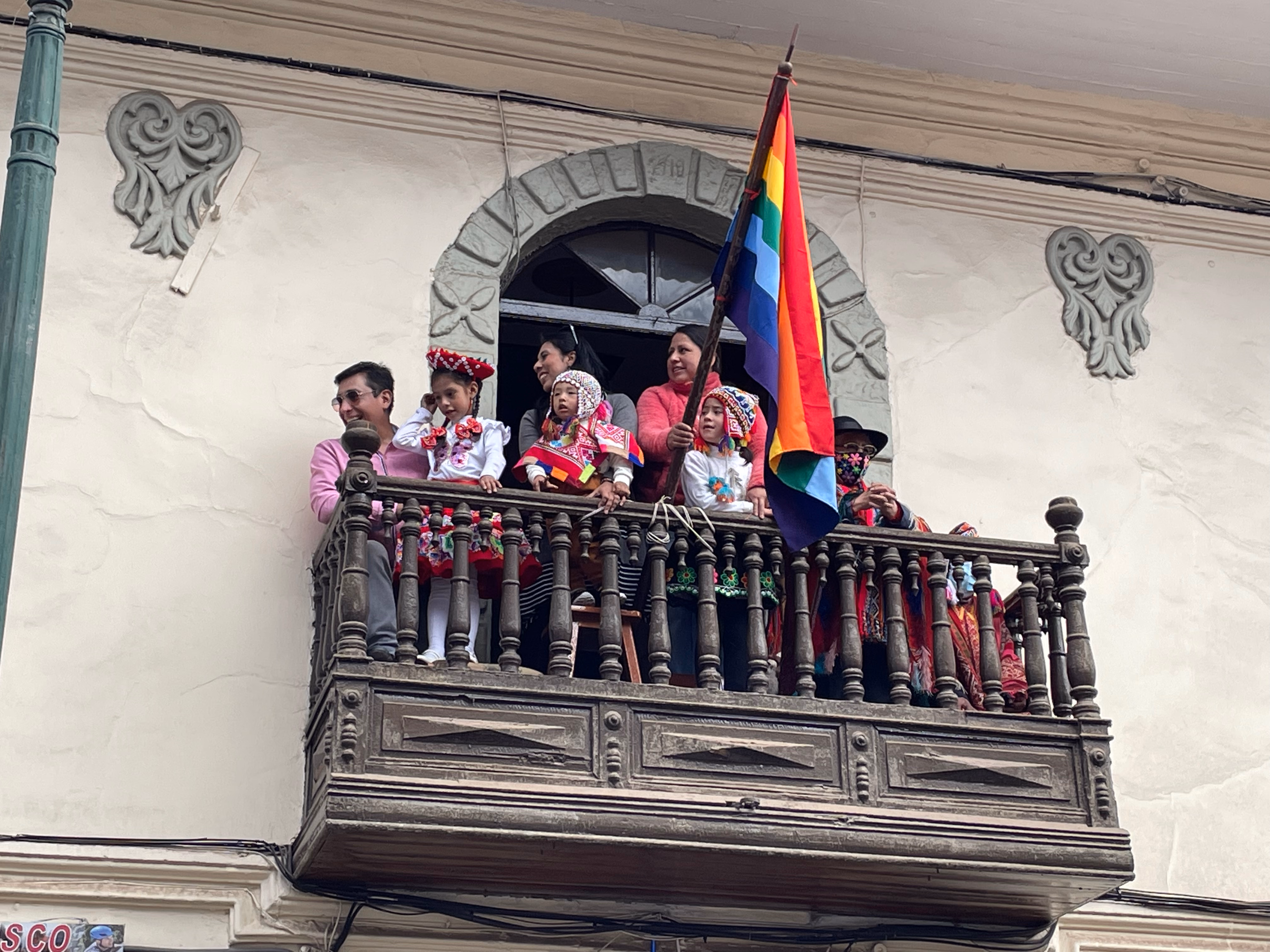 A family watches from a carved wooden balcony — a woman holds the seven-stripe Cusco flag on a pole, children in embroidered skirts and chullos leaning over the balustrade.