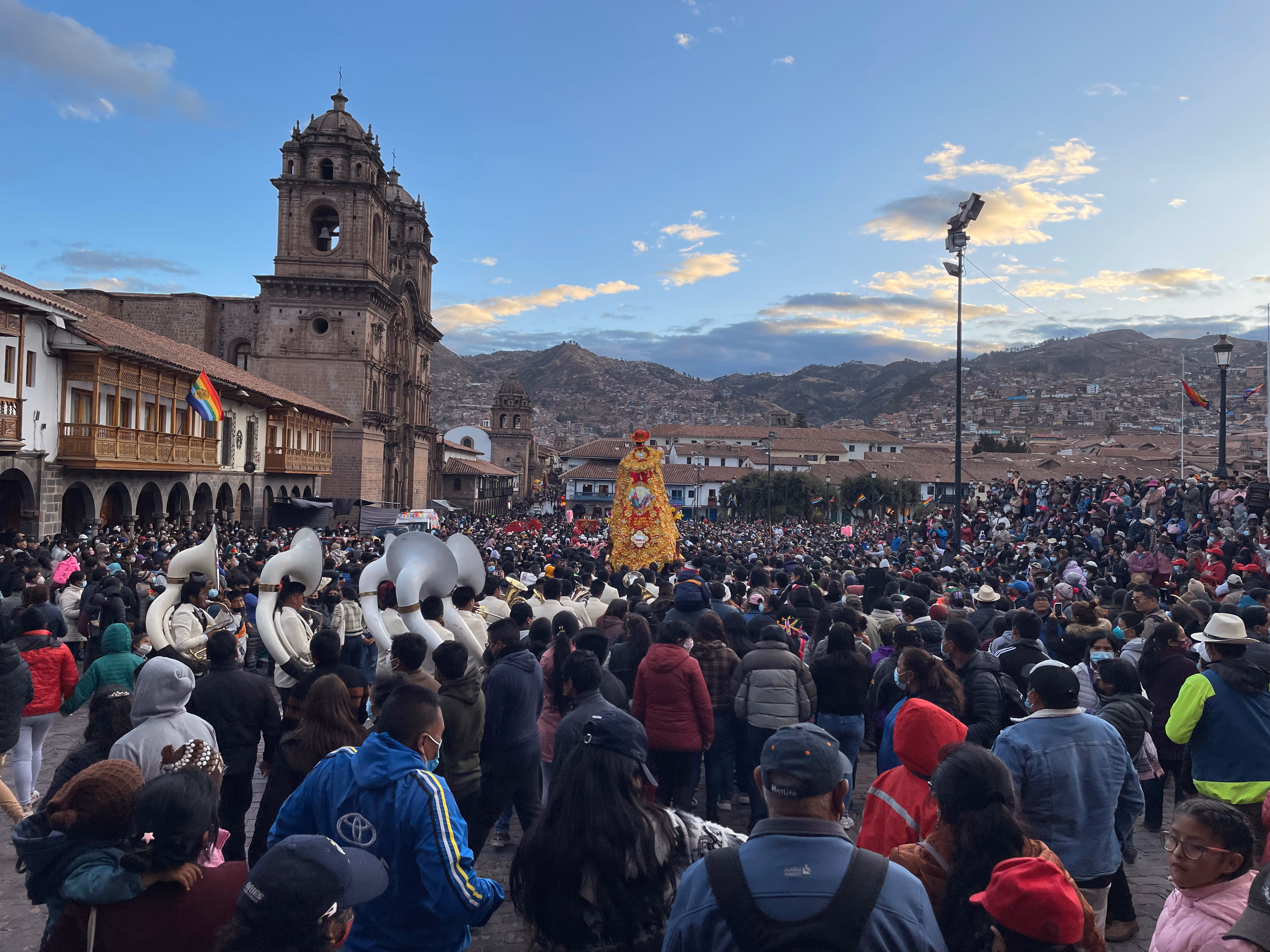 Corpus Christi in the Plaza de Armas at dusk — a golden saint's float carried above the crowd, a brass band with white sousaphones on the left, the Compañía de Jesús lit by the setting sun.
