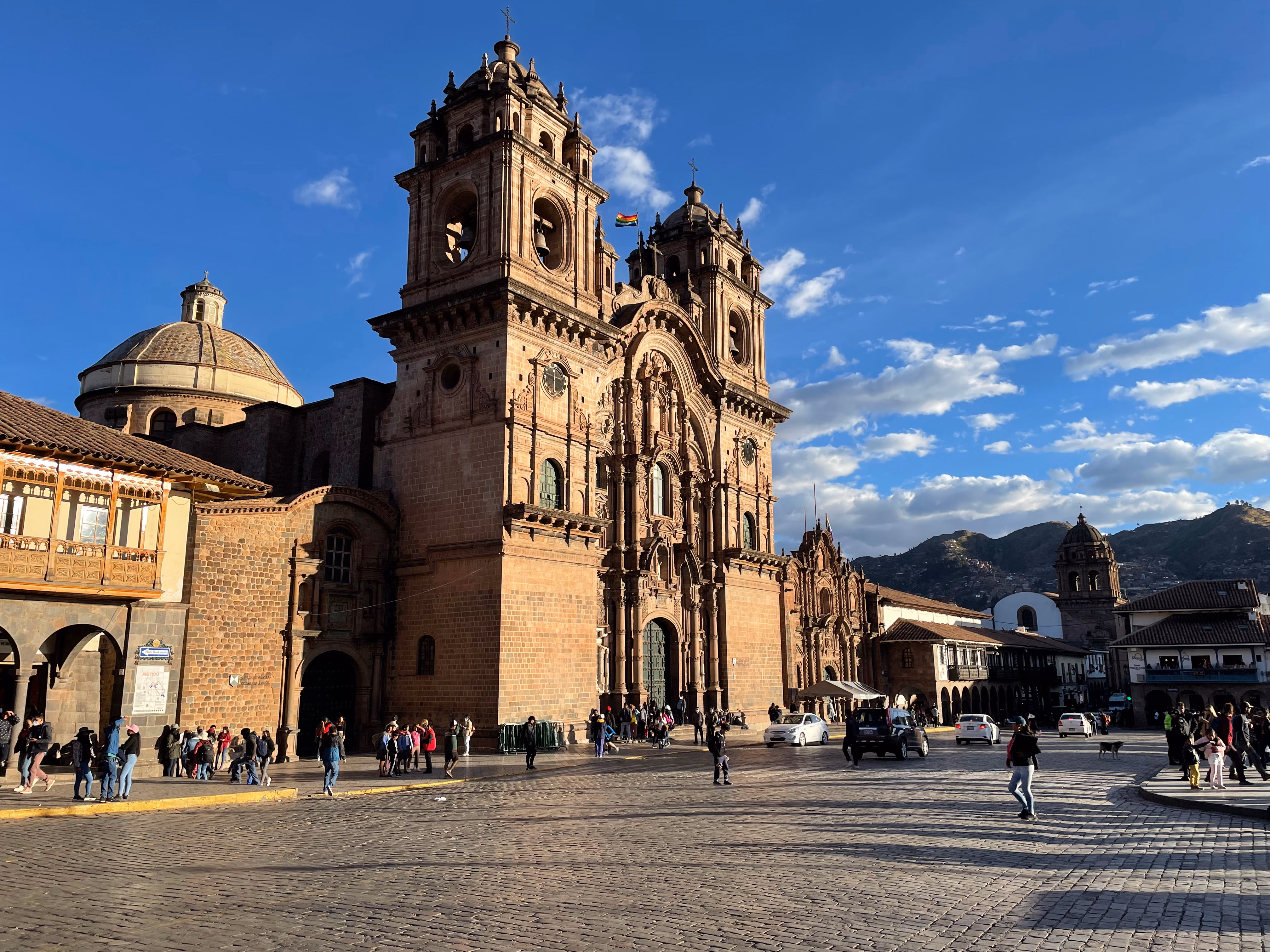 The Church of the Society of Jesus from above — carved brown stone facade, Cusco flag between the bell towers, long shadows stretching across the cobblestones of the main square.