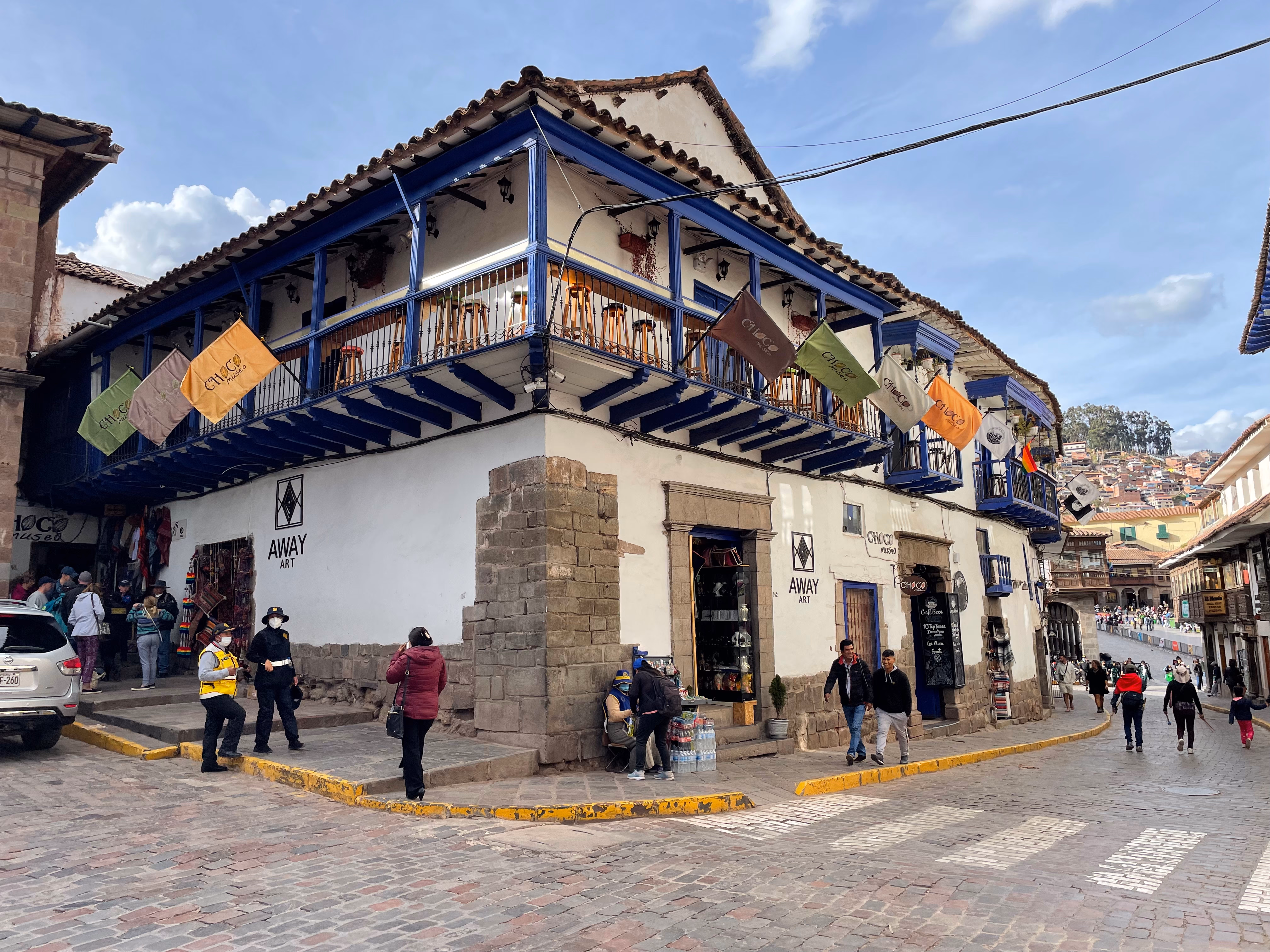 A corner building on Inca stone foundations — white walls, royal-blue wooden balconies draped in festival bunting, cobblestones and terracotta roofs climbing the hillside beyond.