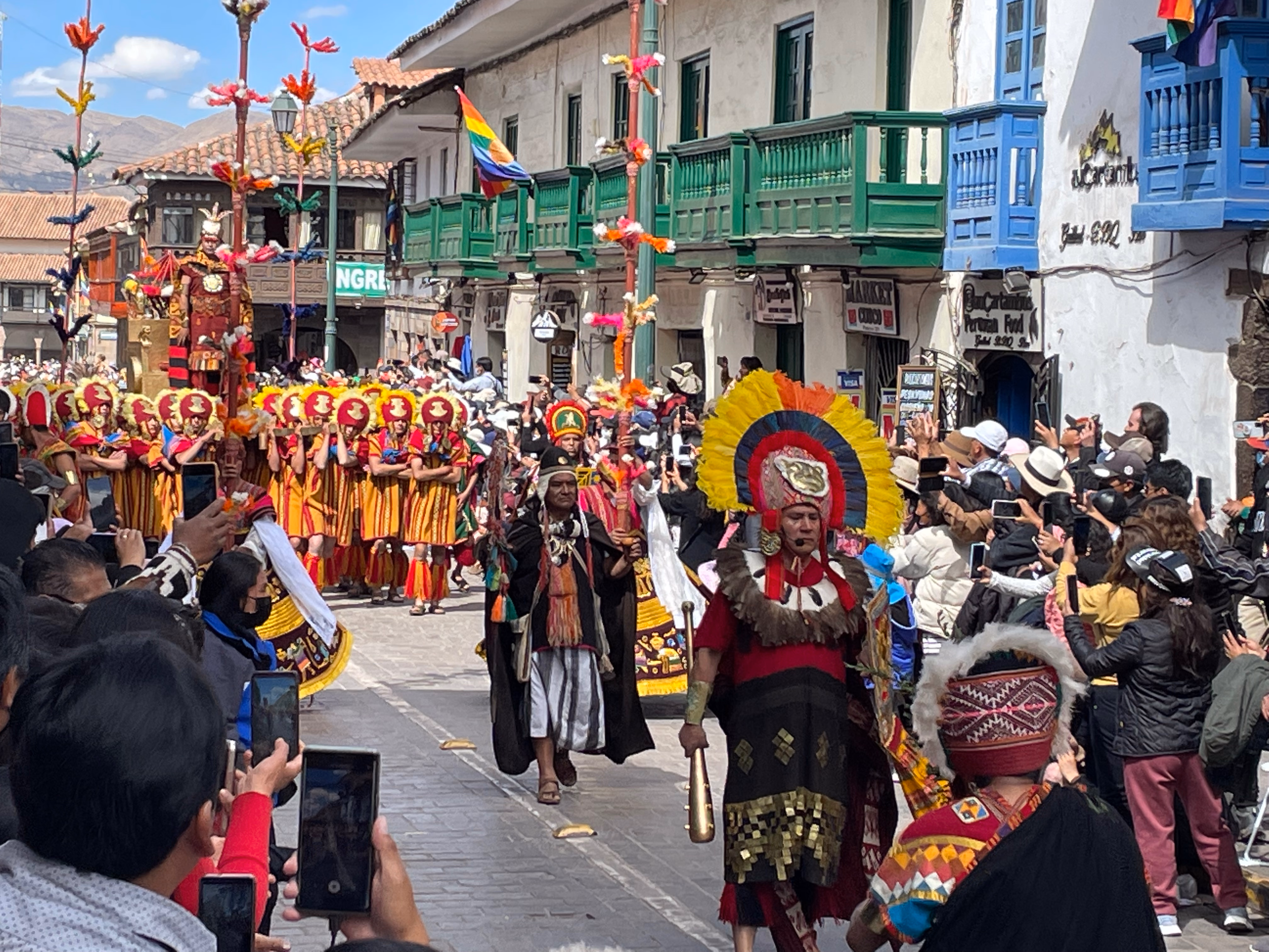 A procession of dancers in red-and-gold tunics and feathered headdresses marches up a cobblestone lane — crowds lining both sides, smartphones raised, green and blue wooden balconies overhead.