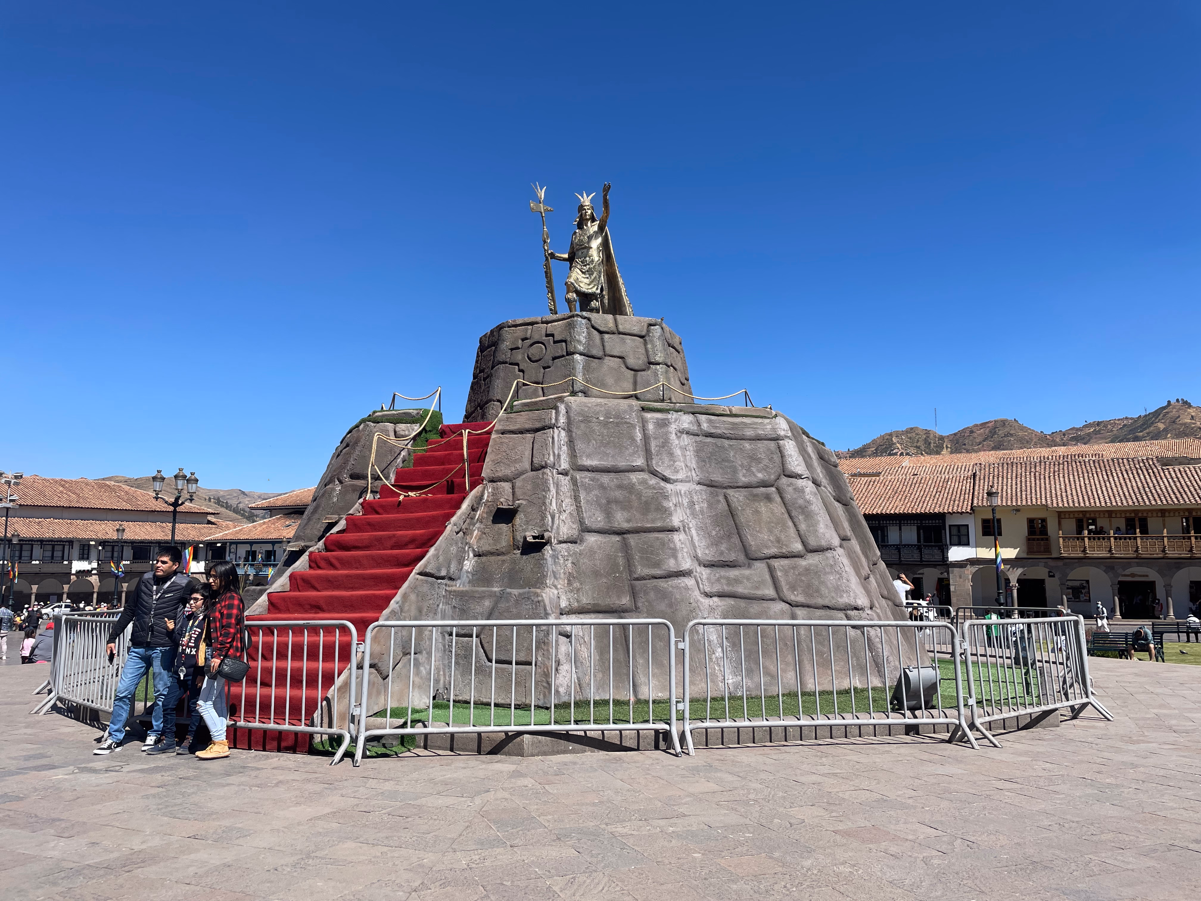 A golden statue of Pachacuti on a tiered stone platform in the Plaza de Armas — red carpet up the stairs, metal barriers around the base, colonial balconies and brown hills beyond.