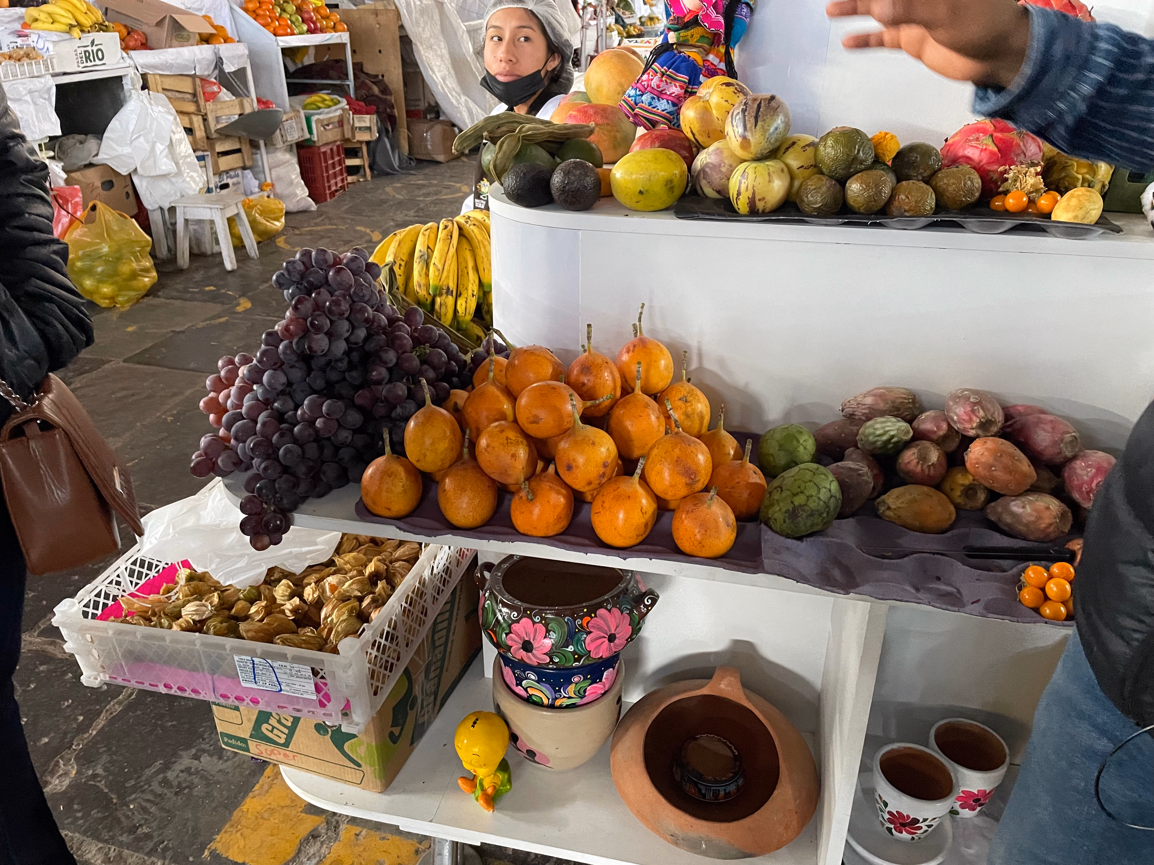Tiered shelves at San Pedro market piled with dragon fruit, cherimoyas, granadillas and prickly pears — painted ceramic bowls and a vendor in a hairnet behind the display.