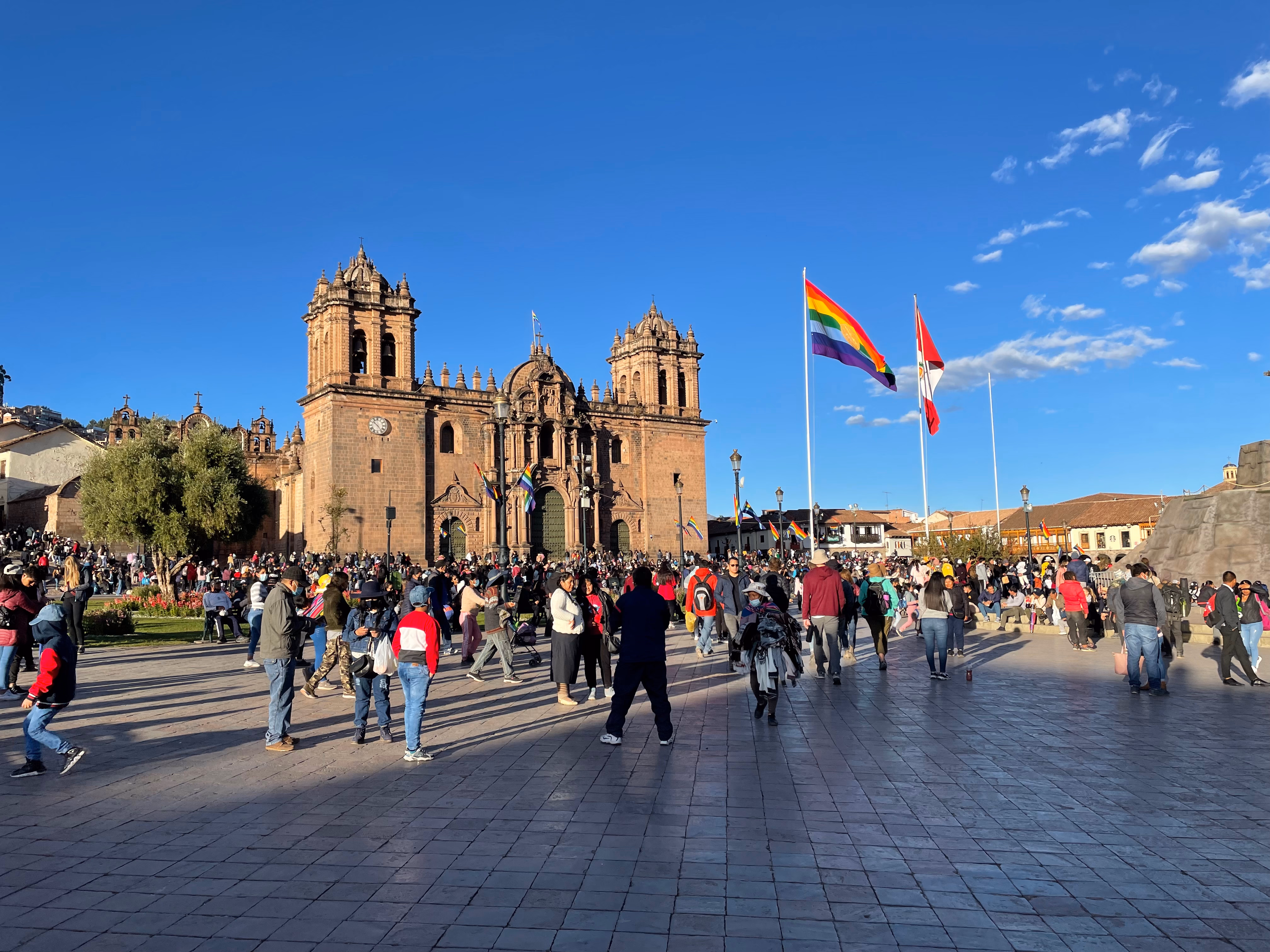 The Plaza de Armas at golden hour — the Cusco rainbow flag and Peruvian tricolour flying above a packed square, the cathedral's twin towers catching the late sun.
