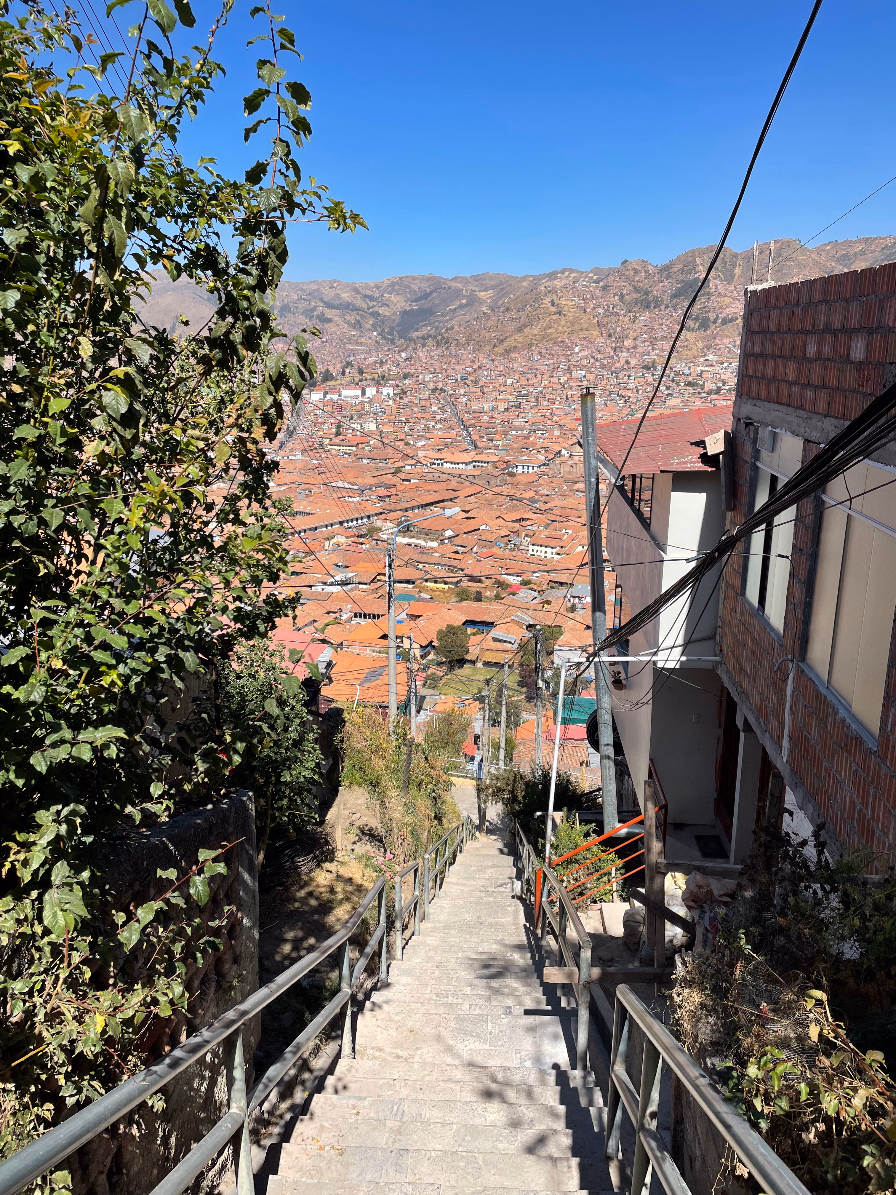 A steep stone stairway with metal handrails plunges toward a sea of terracotta rooftops — the city filling the valley floor, dry brown mountains rising behind.