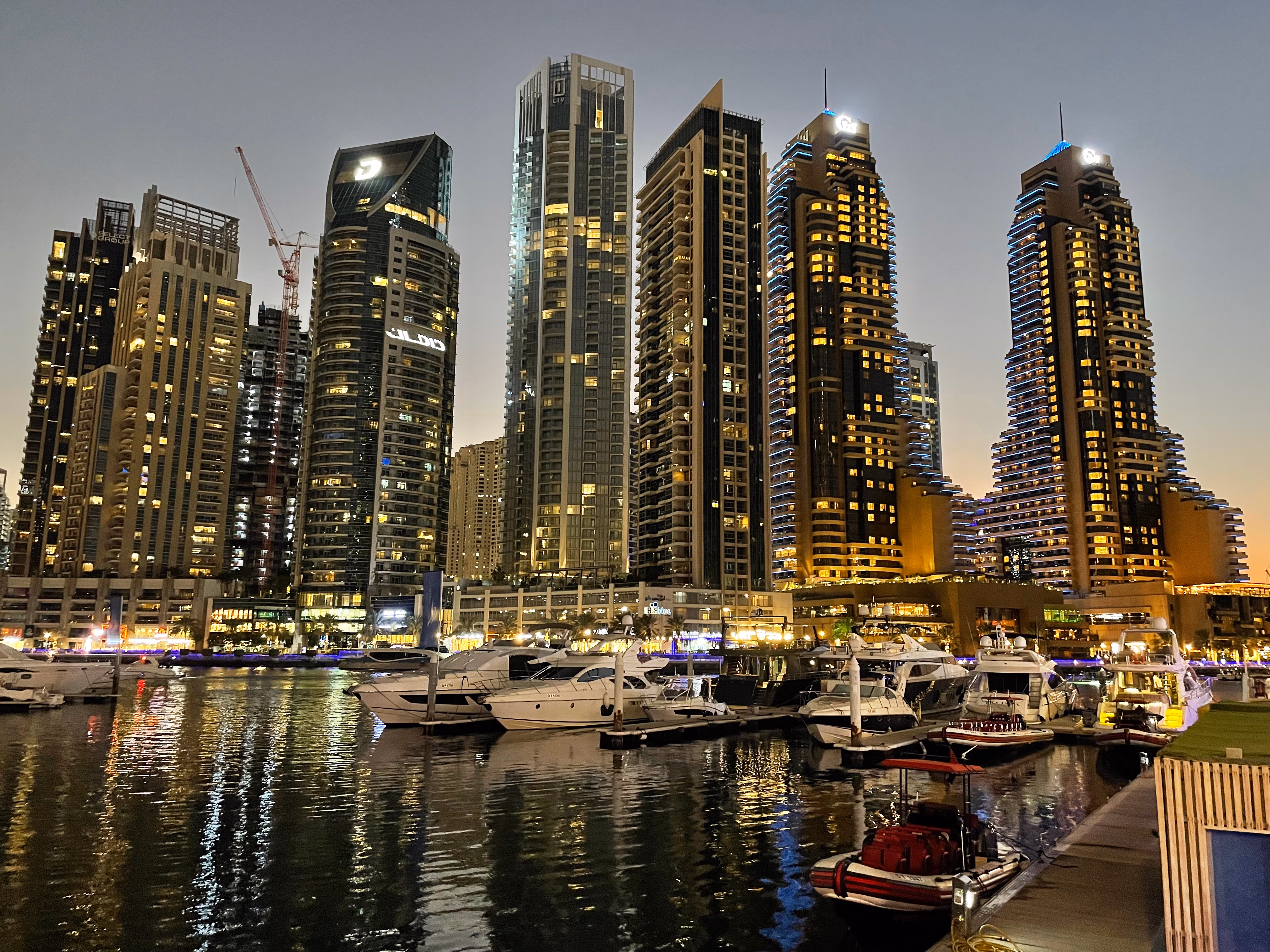 Dubai Marina at dusk — yachts moored below a wall of lit towers, their reflections rippling across the dark water.