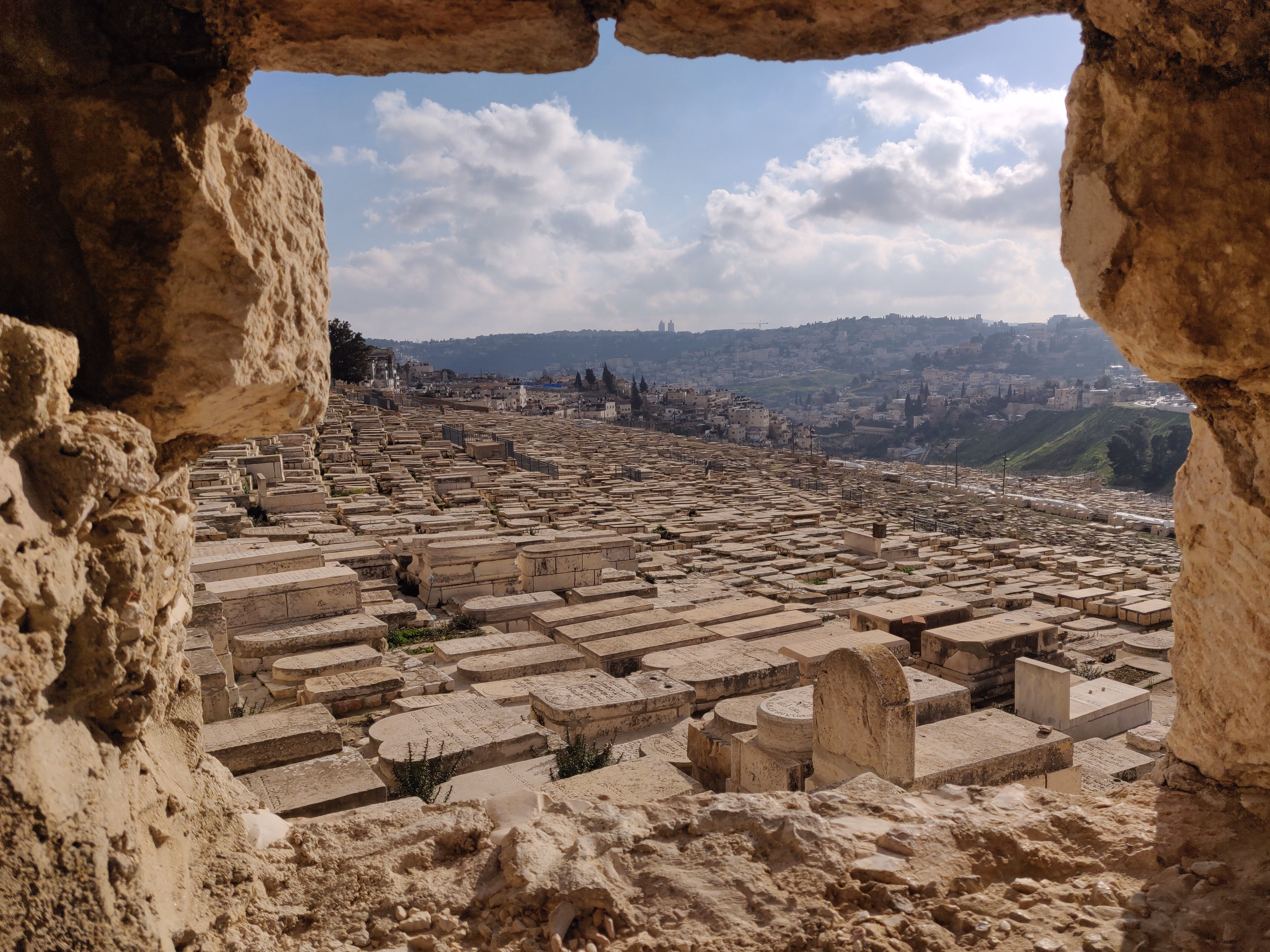 The Mount of Olives cemetery framed through a breach in an ancient wall — rows of pale stone tombs stretching across the hillside toward the city beyond.