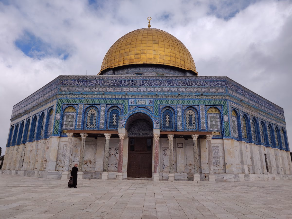 The Dome of the Rock on the Temple Mount — blue and turquoise tilework, white marble and a golden dome catching the light on an open stone plaza.