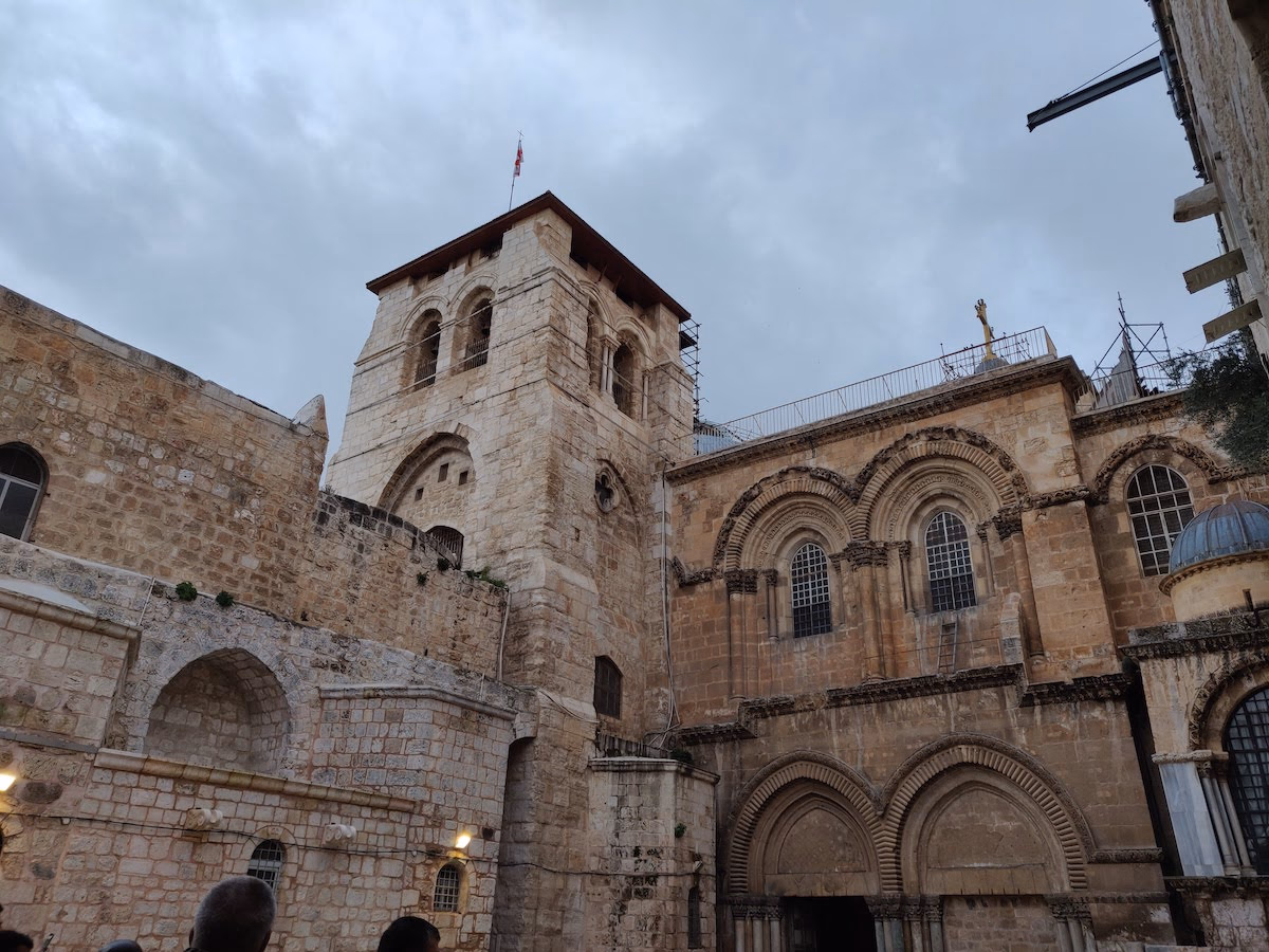 The Church of the Holy Sepulchre under a grey sky — weathered limestone, a square bell tower and ornate arched windows above the Christian Quarter.