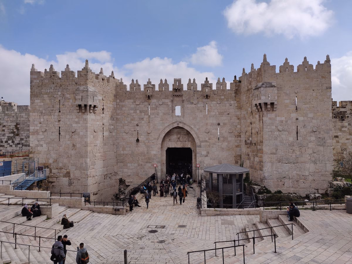 Damascus Gate on a bright afternoon — Ottoman battlements and a pointed arch above a wide stone plaza where locals and visitors gather on the steps.