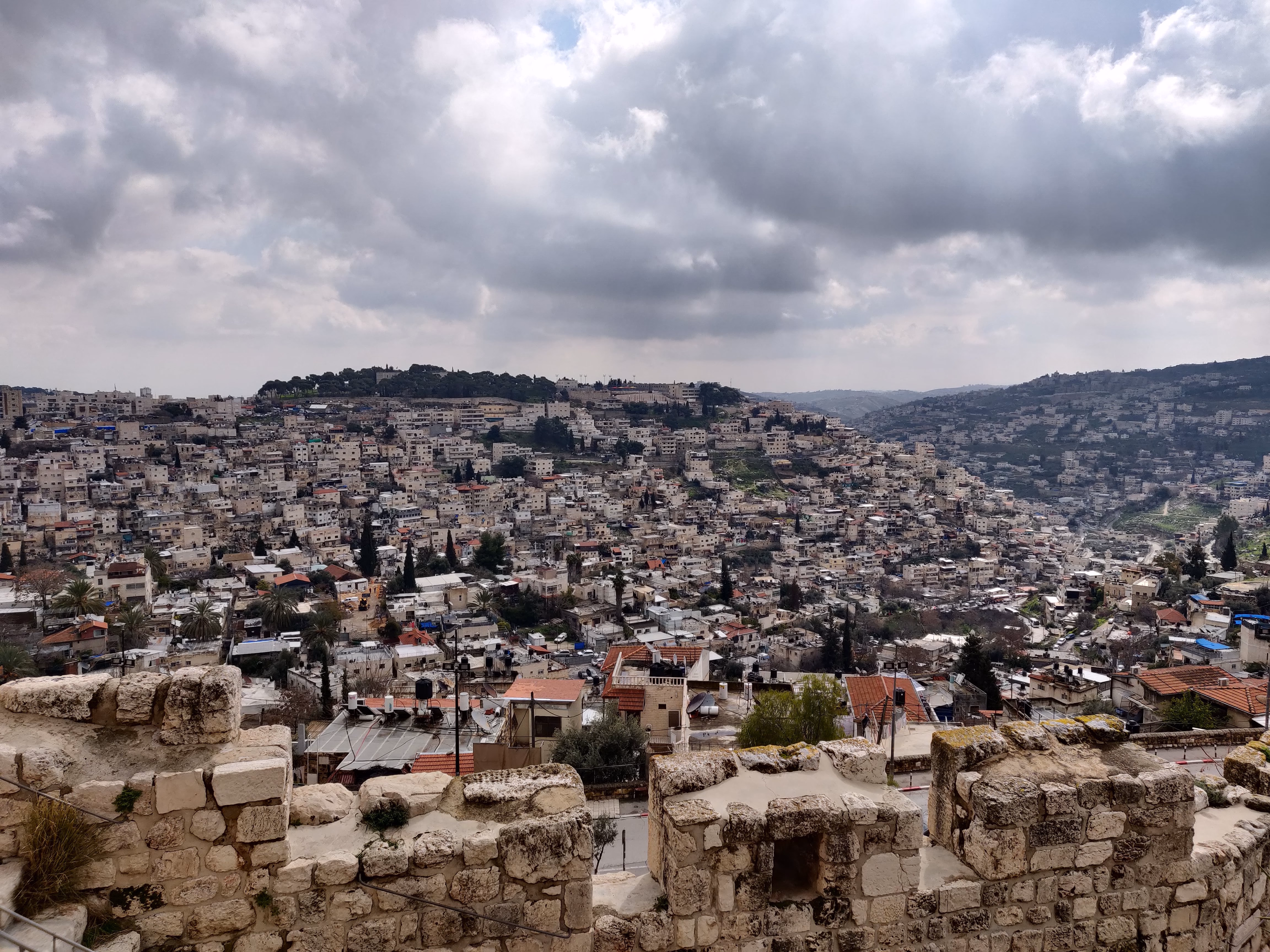 A view from the ramparts over densely packed stone rooftops — water tanks, satellite dishes and cypress trees climbing the hills under heavy grey clouds.