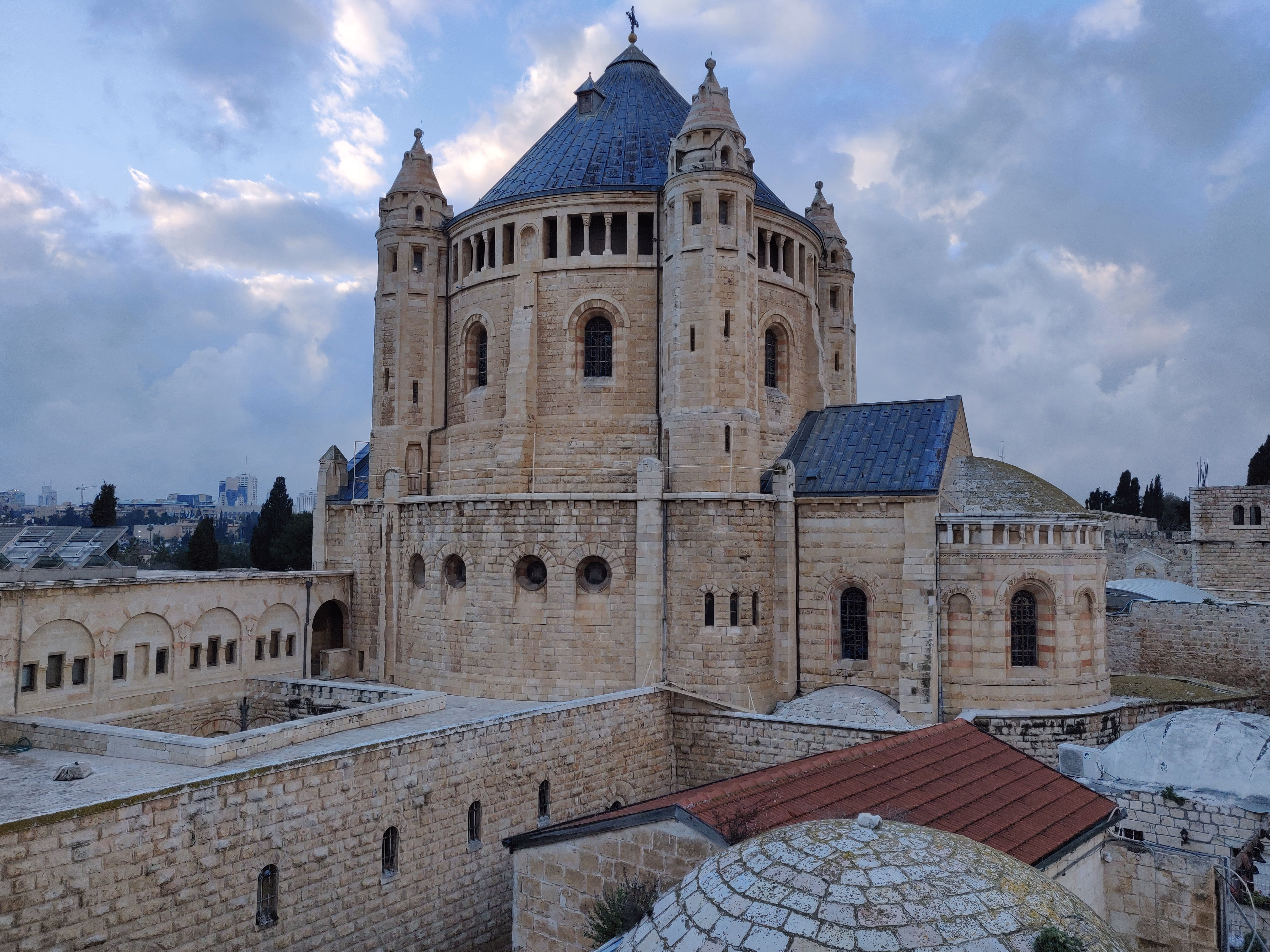 The Abbey of the Dormition on Mount Zion — a round stone church with a dark conical roof and four corner towers, the modern skyline faint on the horizon.