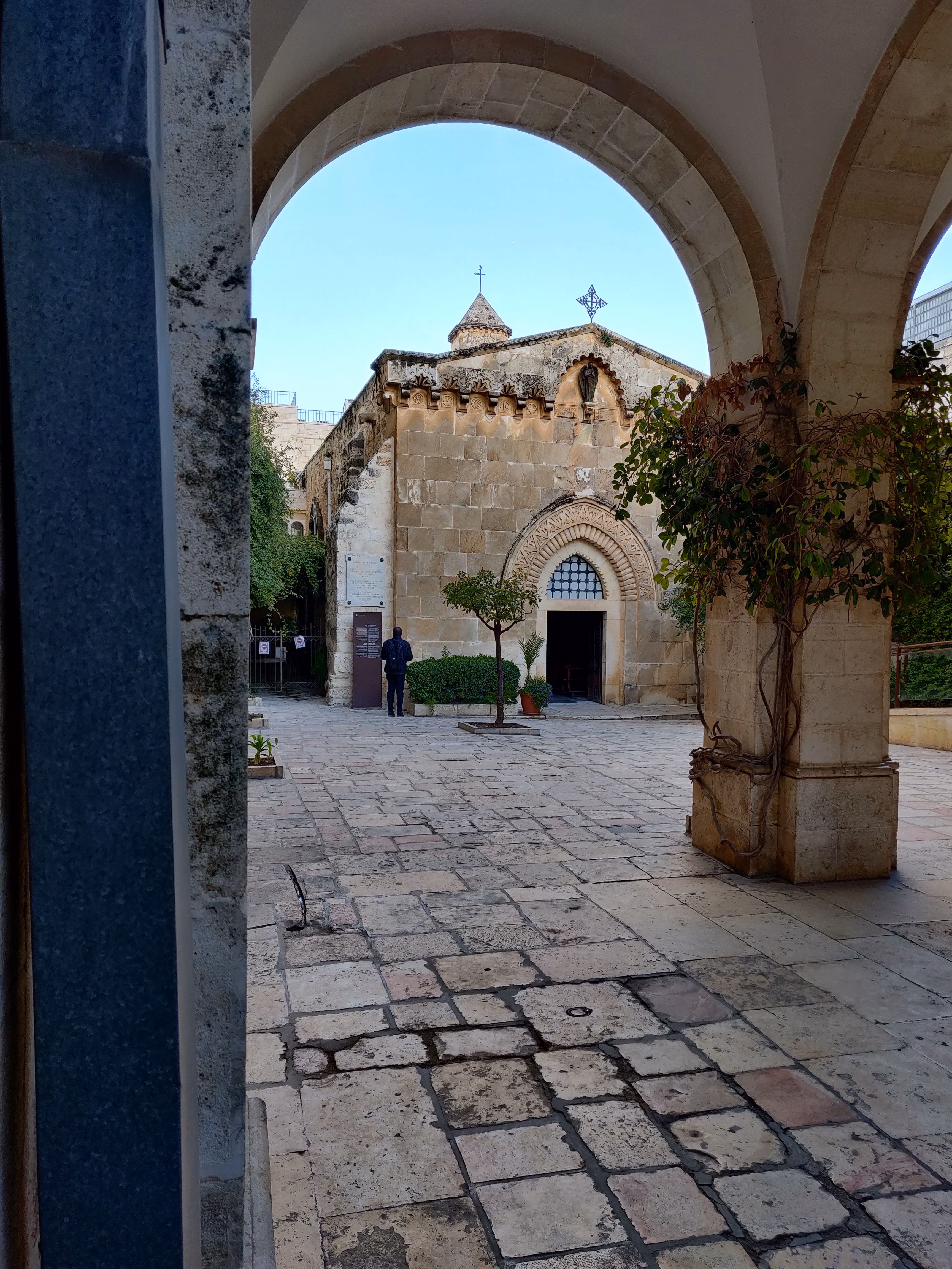 A courtyard on the Via Dolorosa seen through a stone arch — chevron-carved doorway, crosses on the roofline and worn paving stones in the sun.