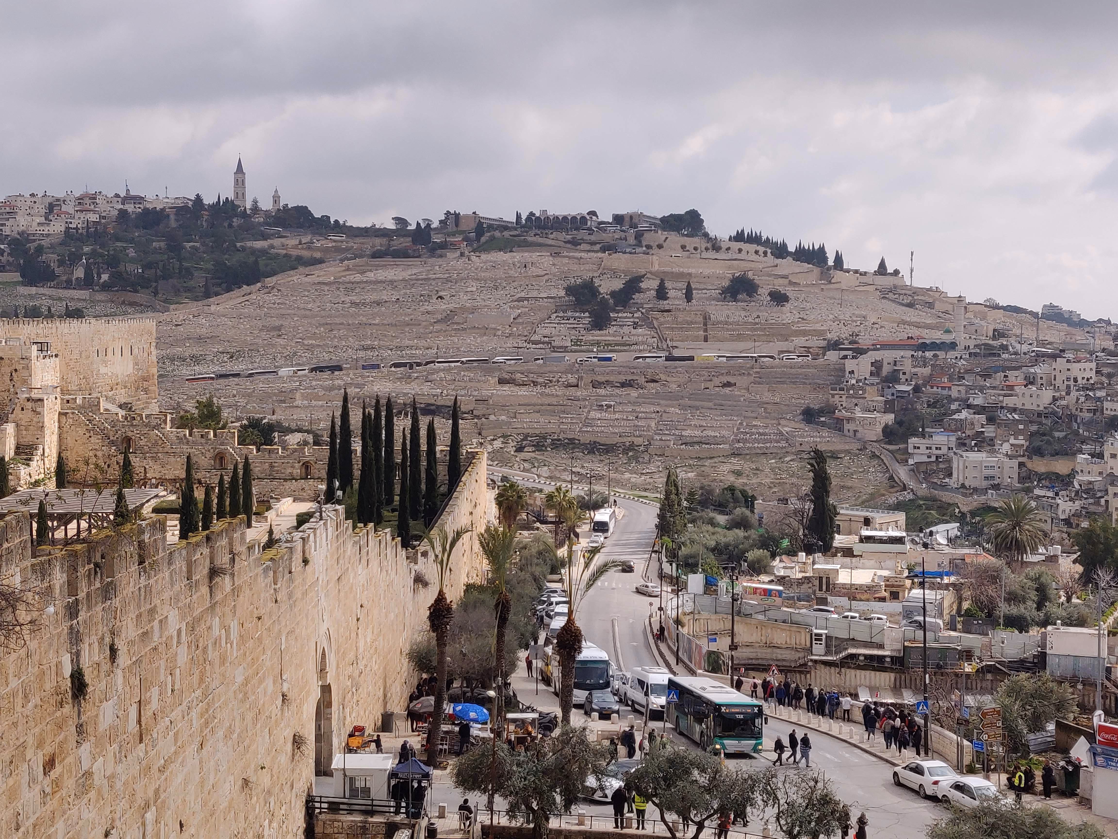 The Old City walls above a busy road — crenellated limestone on the left, the Mount of Olives cemetery blanketing the hillside beyond.