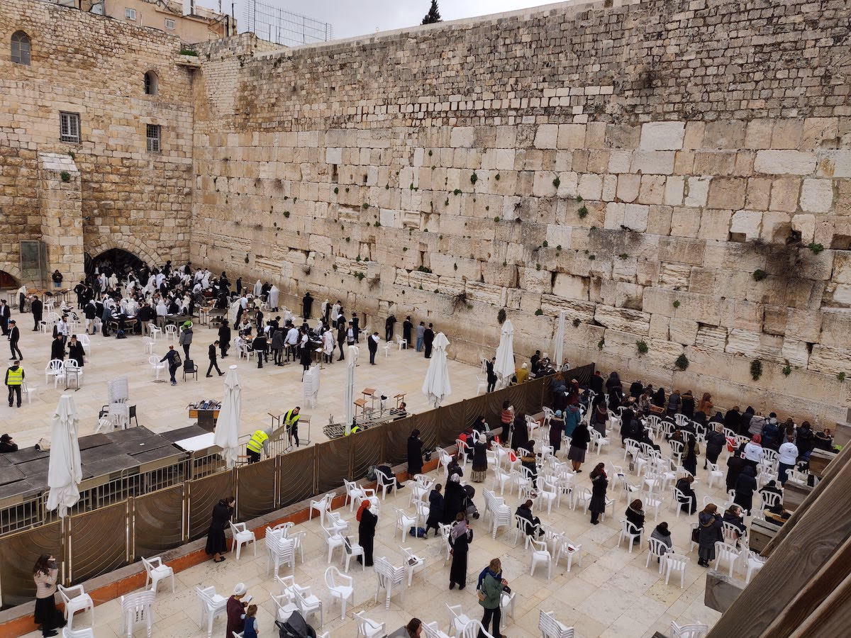 The Western Wall plaza from above — worshippers at the ancient limestone face, white chairs and a mechitza dividing the men's and women's sections.