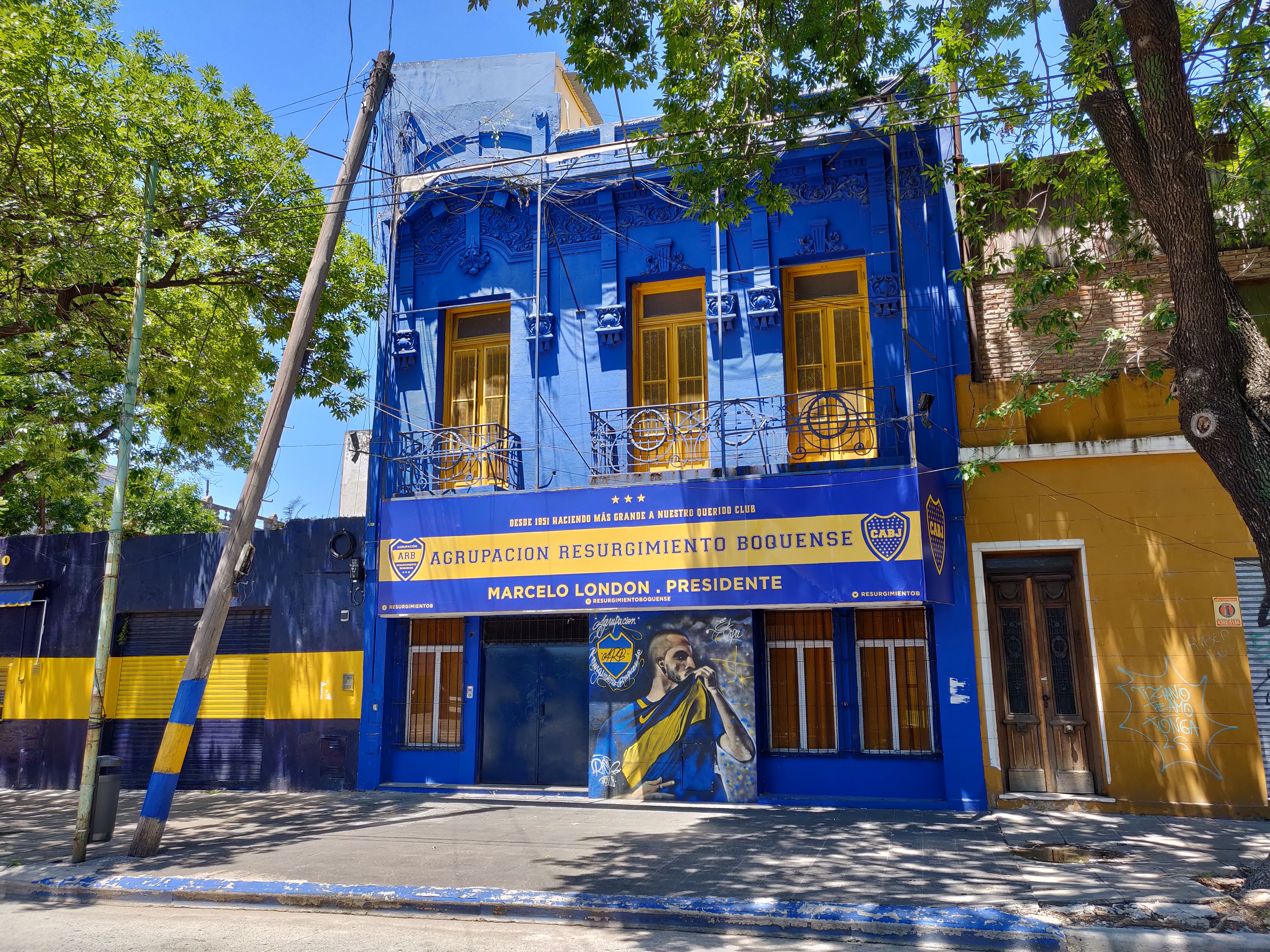 A deep-blue townhouse draped in Boca Juniors banners — yellow shutters, the CABJ crest on a mural and a player bowing to kiss the club shield.
