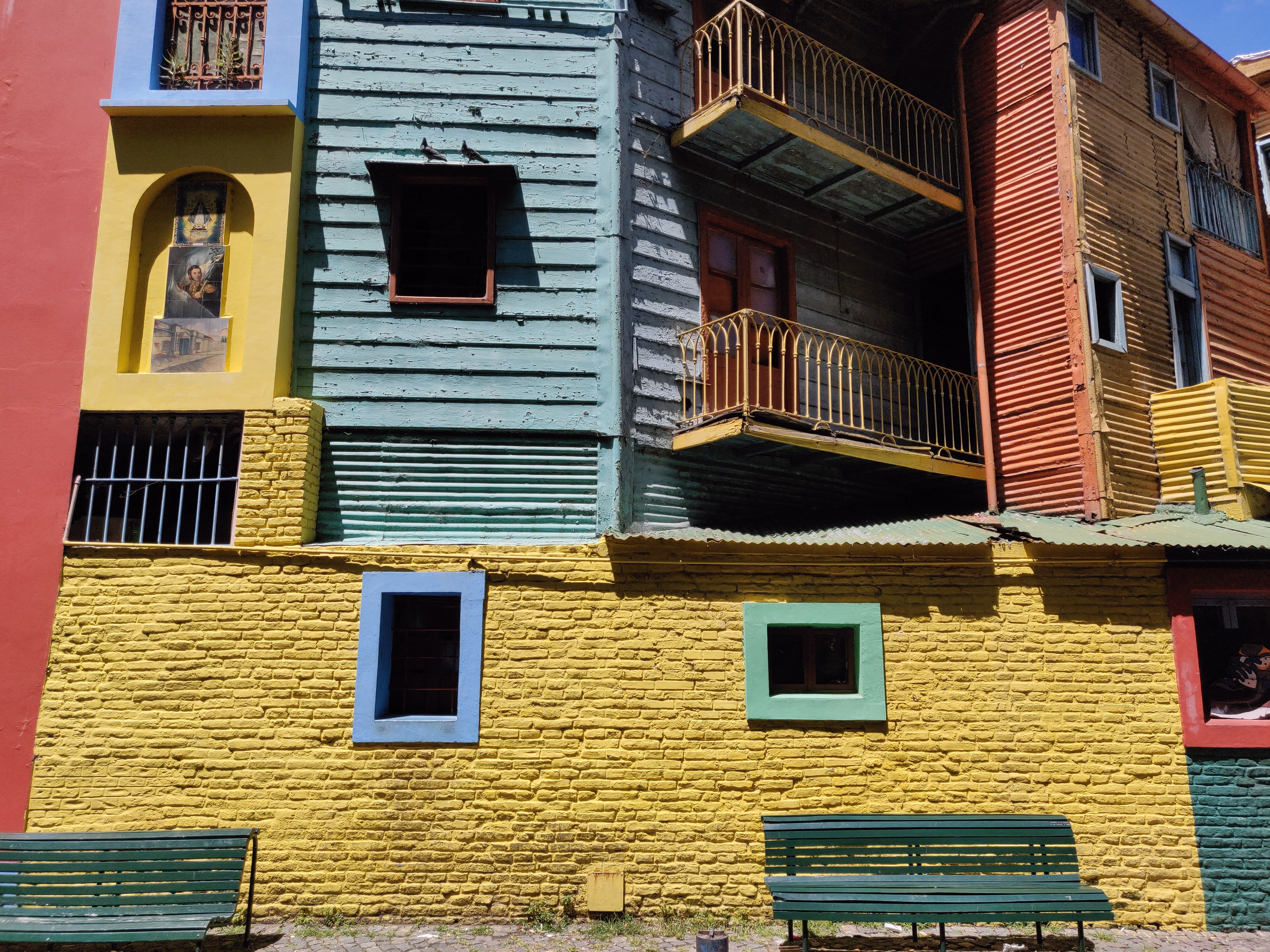 Patchwork corrugated-metal walls in yellow, orange, blue and red — painted window frames, curved iron balconies and green park benches at the base.