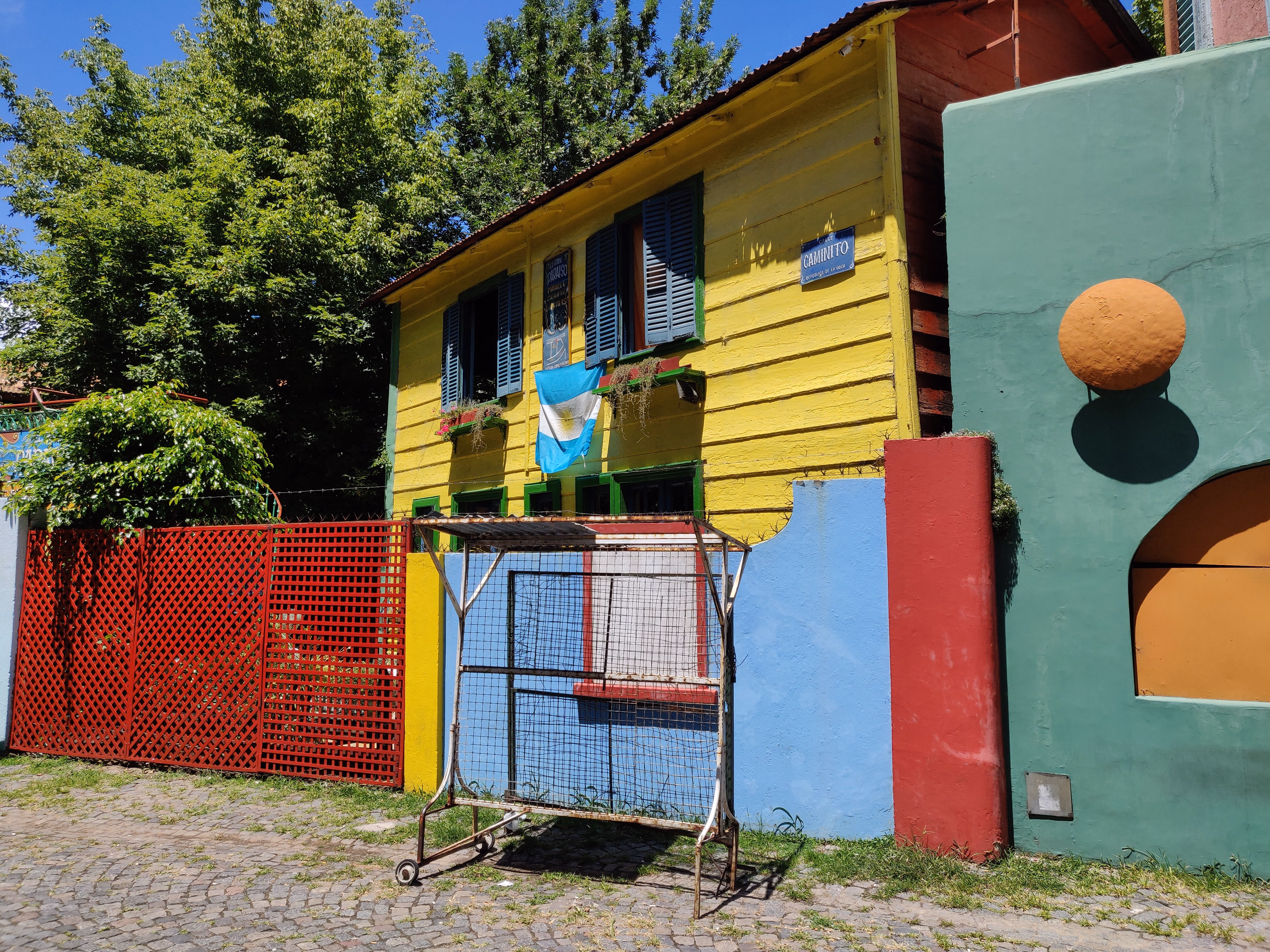 A Caminito street sign on corrugated yellow siding, an Argentine flag draped over the window and a football goal frame standing on the cobbles.