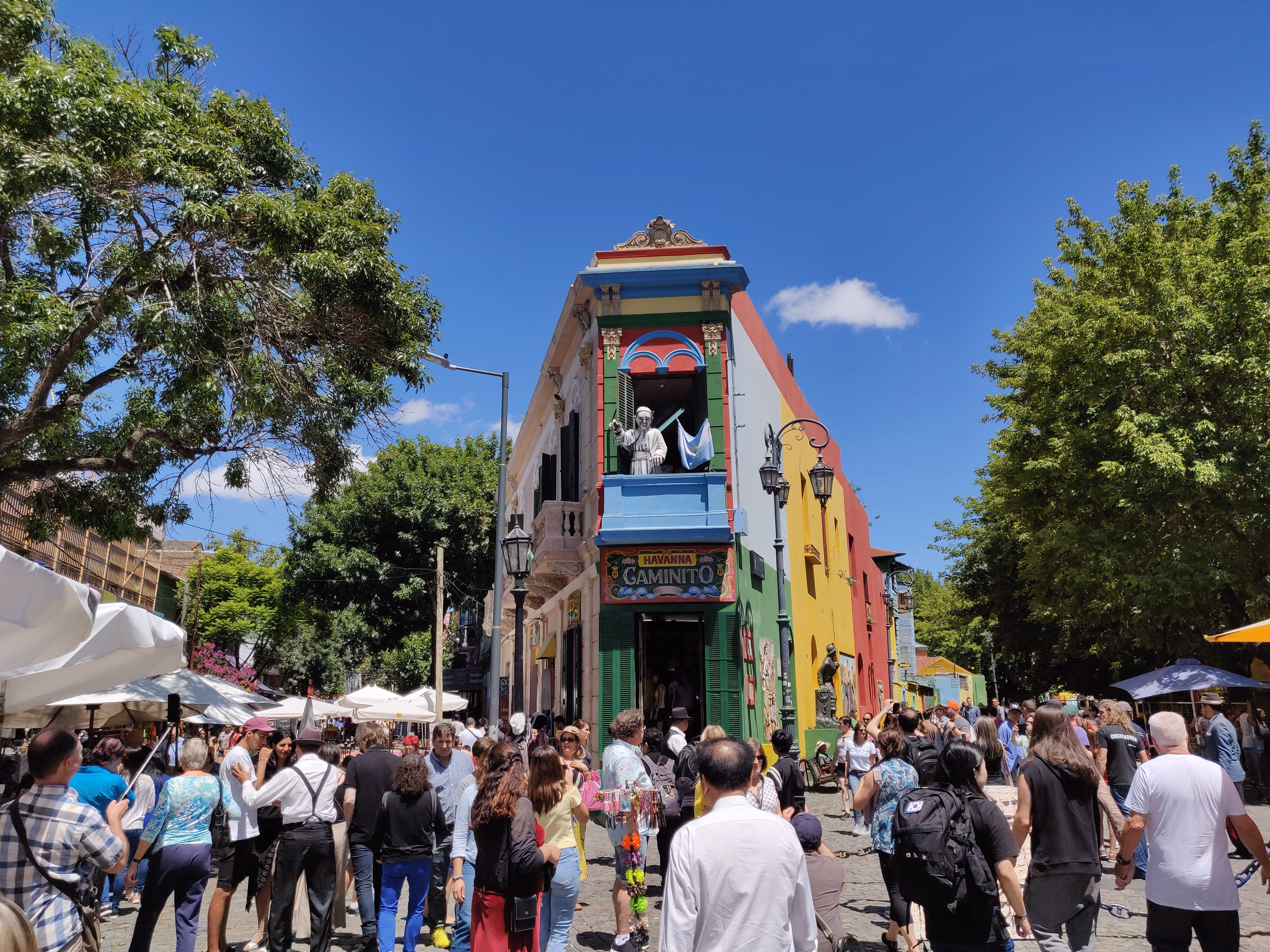 The Havanna Caminito corner on a busy afternoon — green, yellow and blue facades, a tango relief on the ground floor and tourists filling the cobblestone lane.
