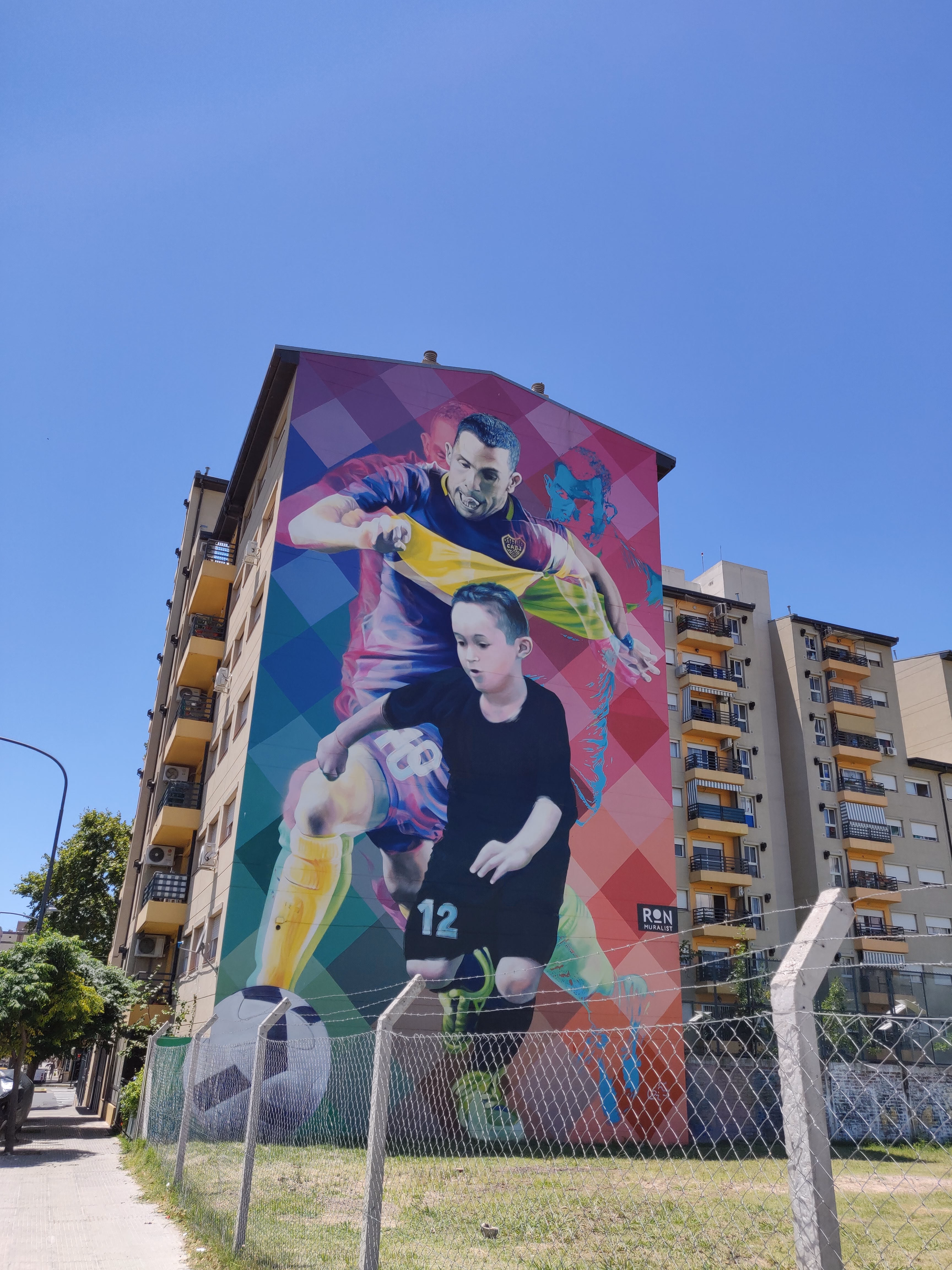 A towering Ron Muralist piece on an apartment block — Carlos Tevez in Boca colours above a boy dribbling, against a harlequin diamond of pink, turquoise and purple.
