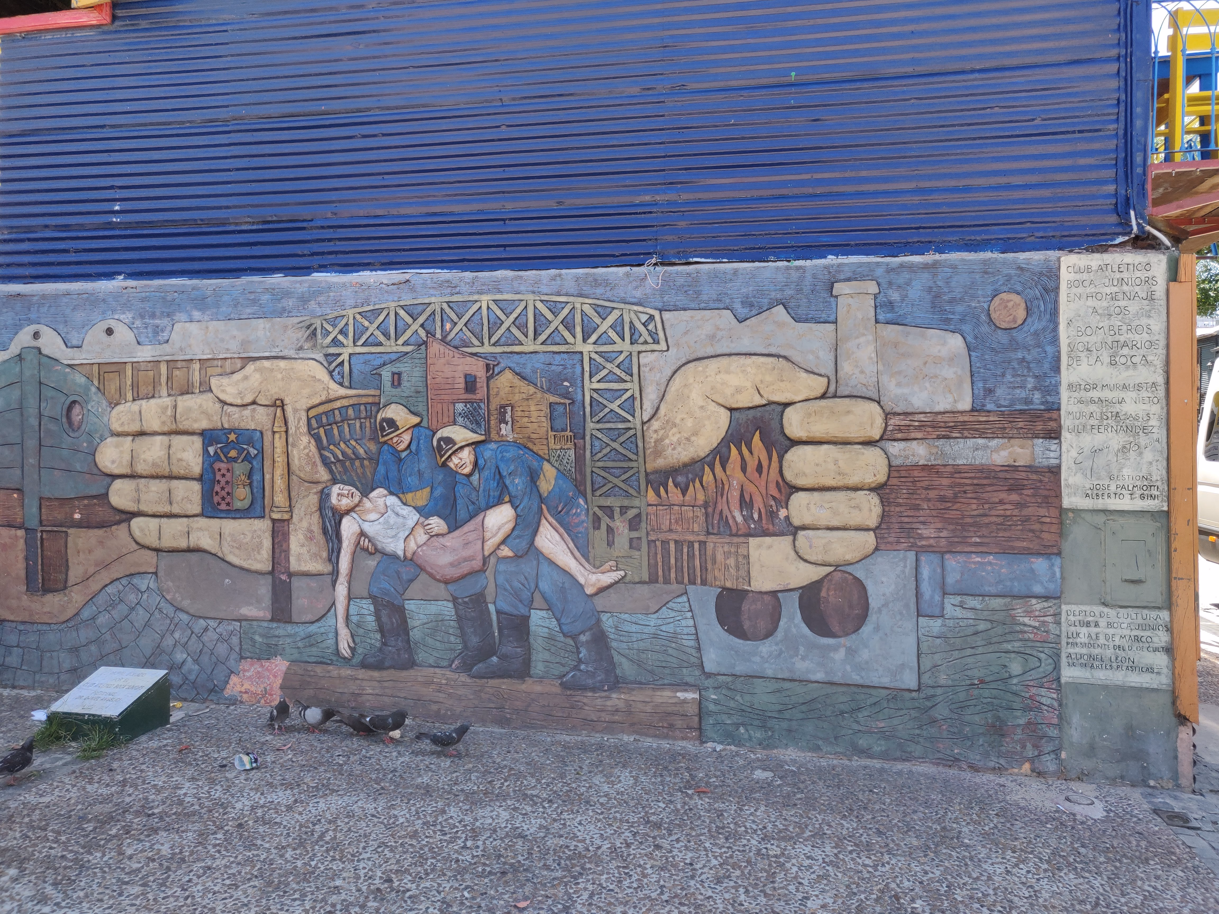 Firefighters carry a woman from a blaze, cradled in giant painted hands — the Boca Juniors crest on the left, a plaque honouring the volunteer bomberos on the right.