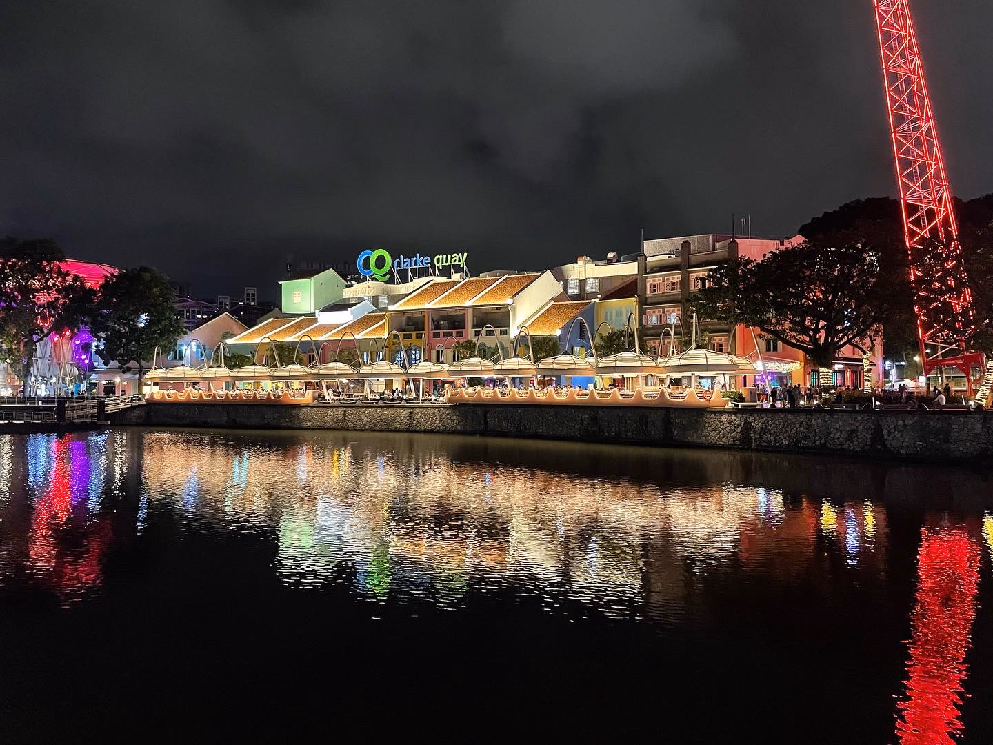 Clarke Quay from the water โ mushroom canopies, the neon CQ sign, and a red lattice ride reflected in long streaks on the river.