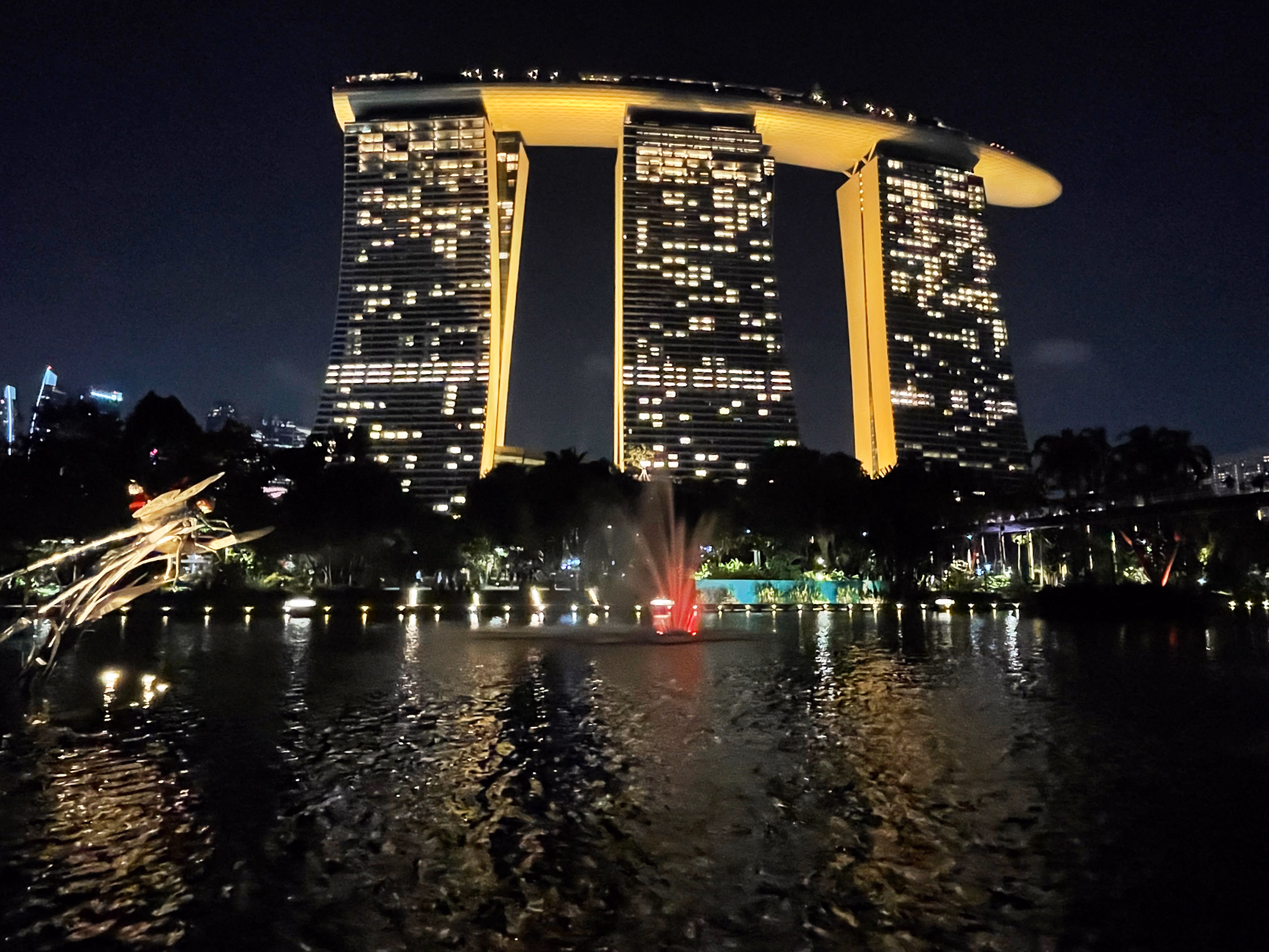 The three towers of Marina Bay Sands from Gardens by the Bay โ a red-lit fountain in the lake, tropical trees in silhouette below the SkyPark.