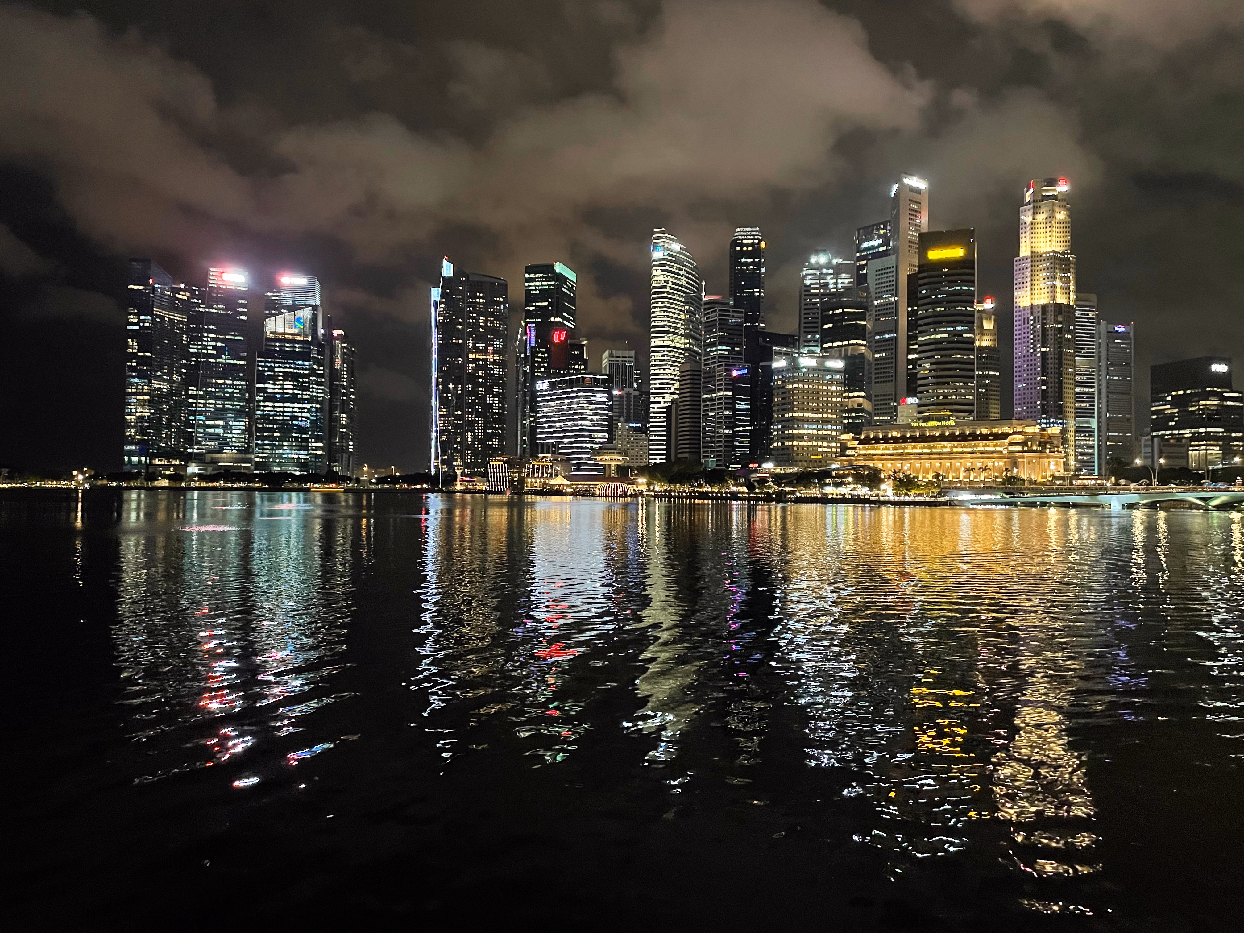 The CBD skyline across Marina Bay โ a wall of lit towers mirrored in long streaks of gold and white on the dark water.