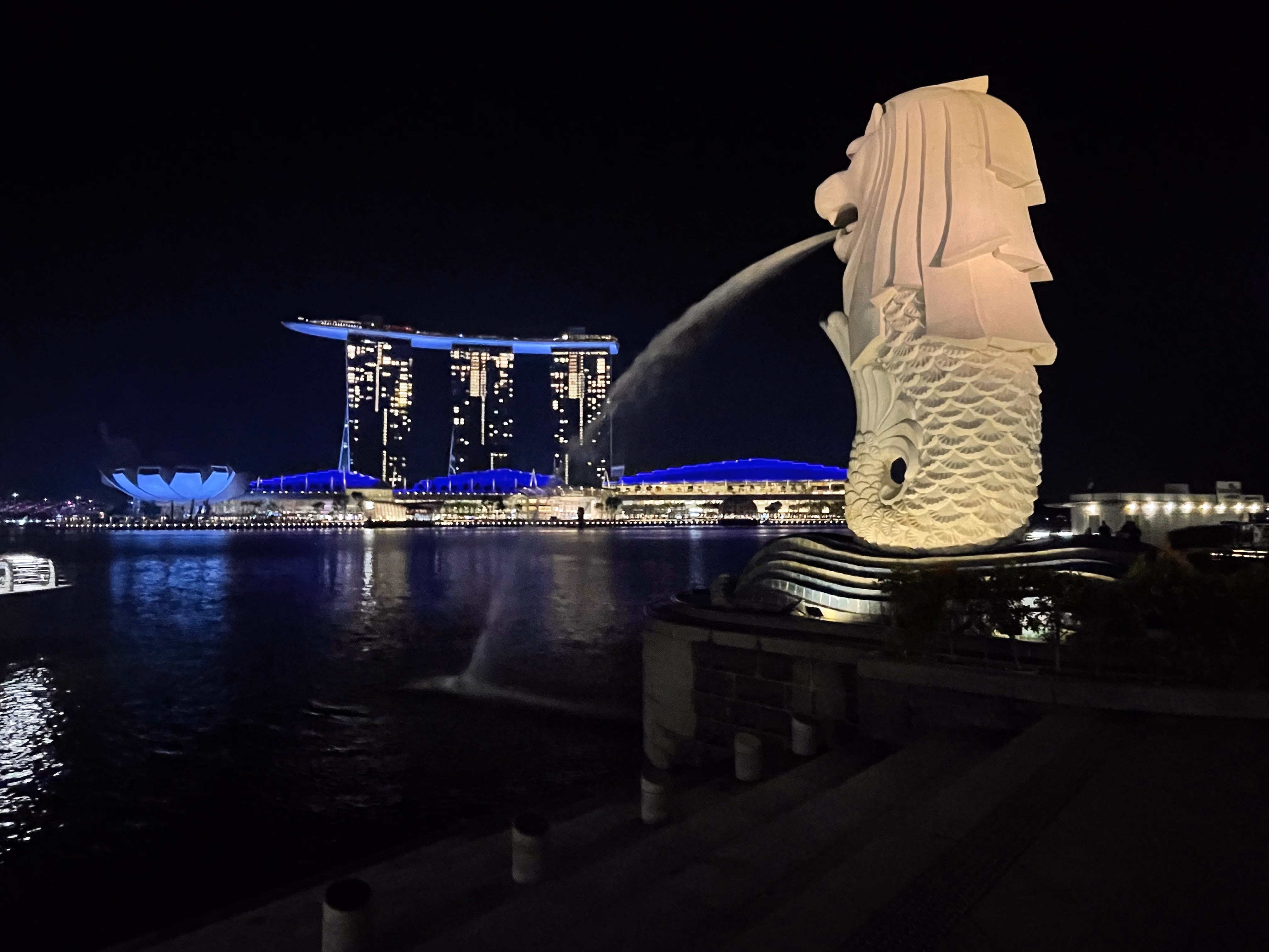 The Merlion on its pier, spouting into the bay โ Marina Bay Sands and the blue ArtScience Museum lined up across the water behind it.