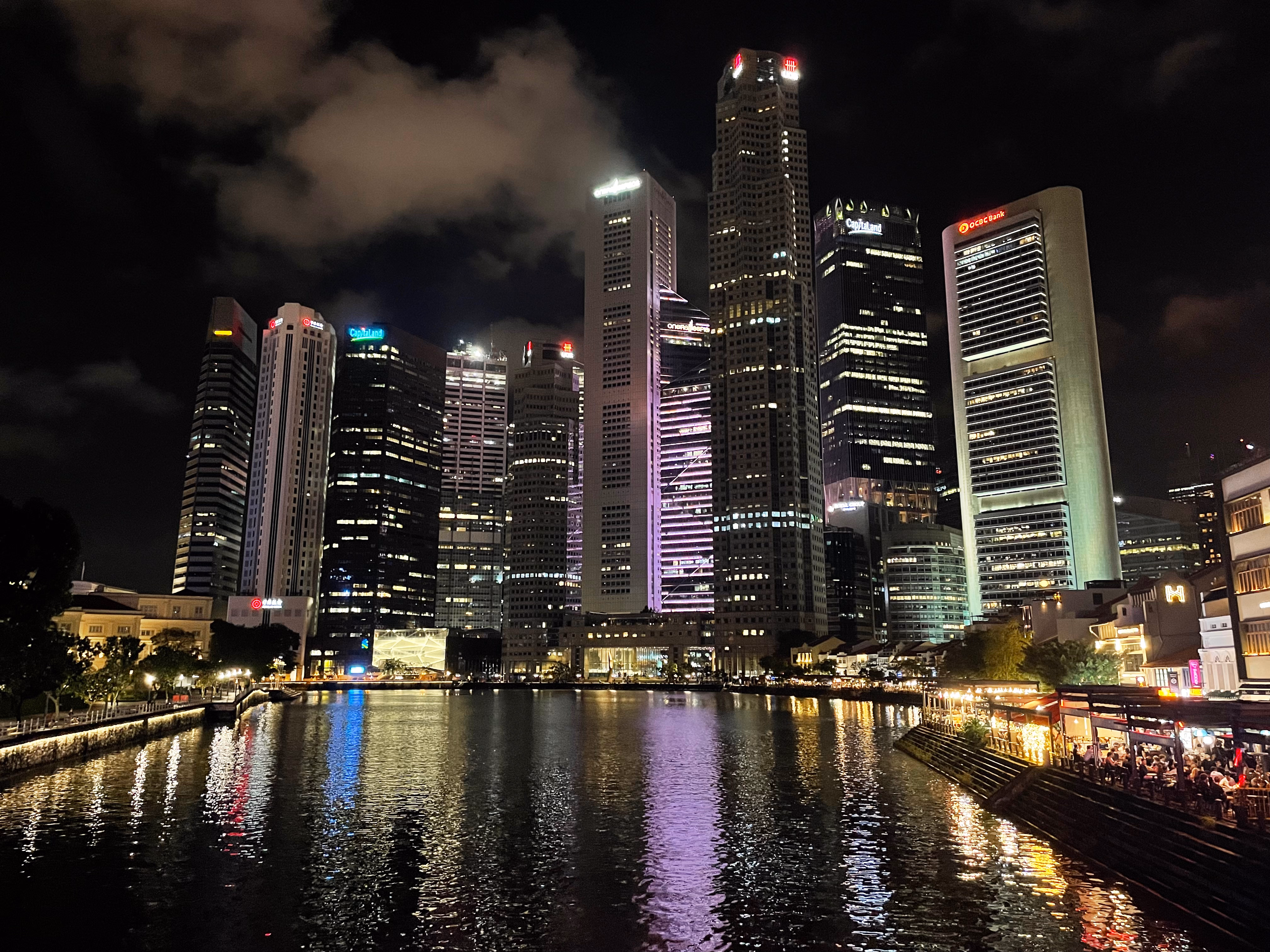 Boat Quay below the CBD โ heritage shophouses and red umbrellas along the bank, skyscrapers rising behind the Singapore River.