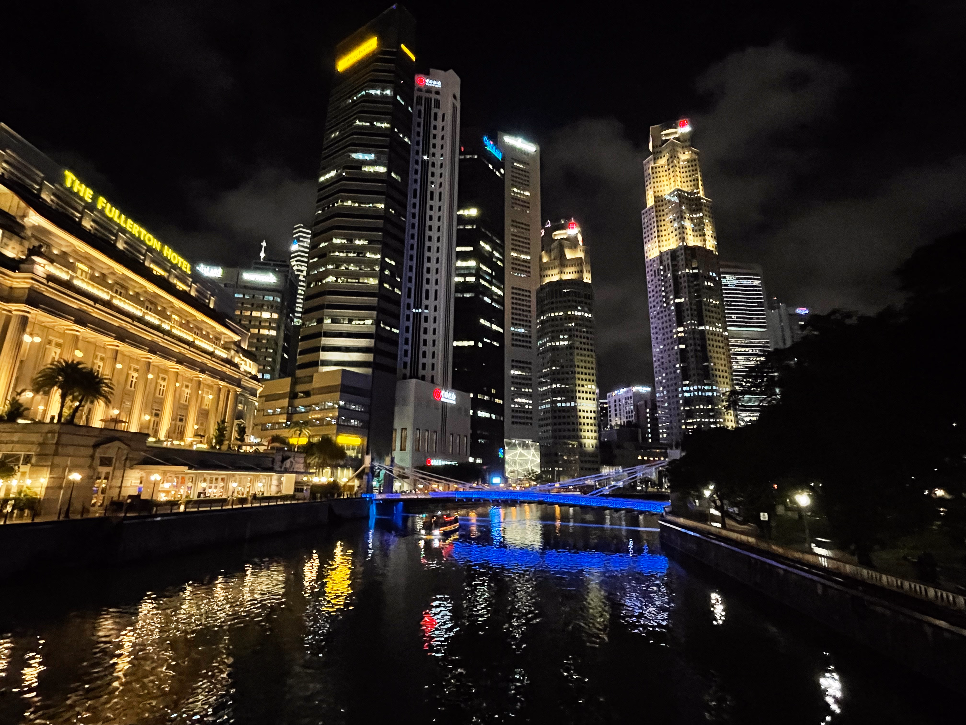 The Fullerton Hotel and a blue-lit bridge on the Singapore River โ colonial colonnades in front of UOB Plaza and the rest of the skyline.