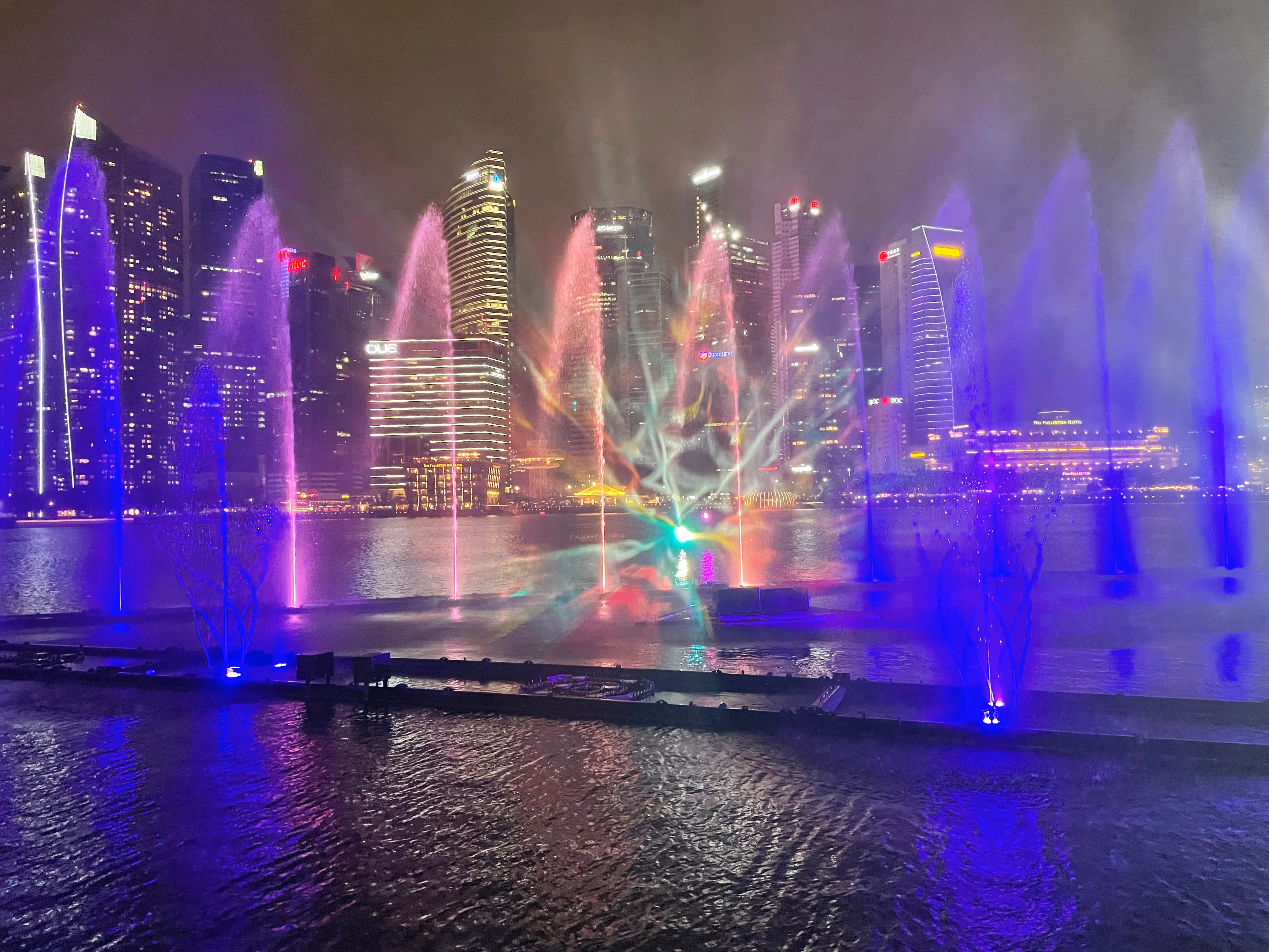Columns of purple and pink water rising from the show platform, a kaleidoscope of colour in the mist and the skyline glowing beyond.