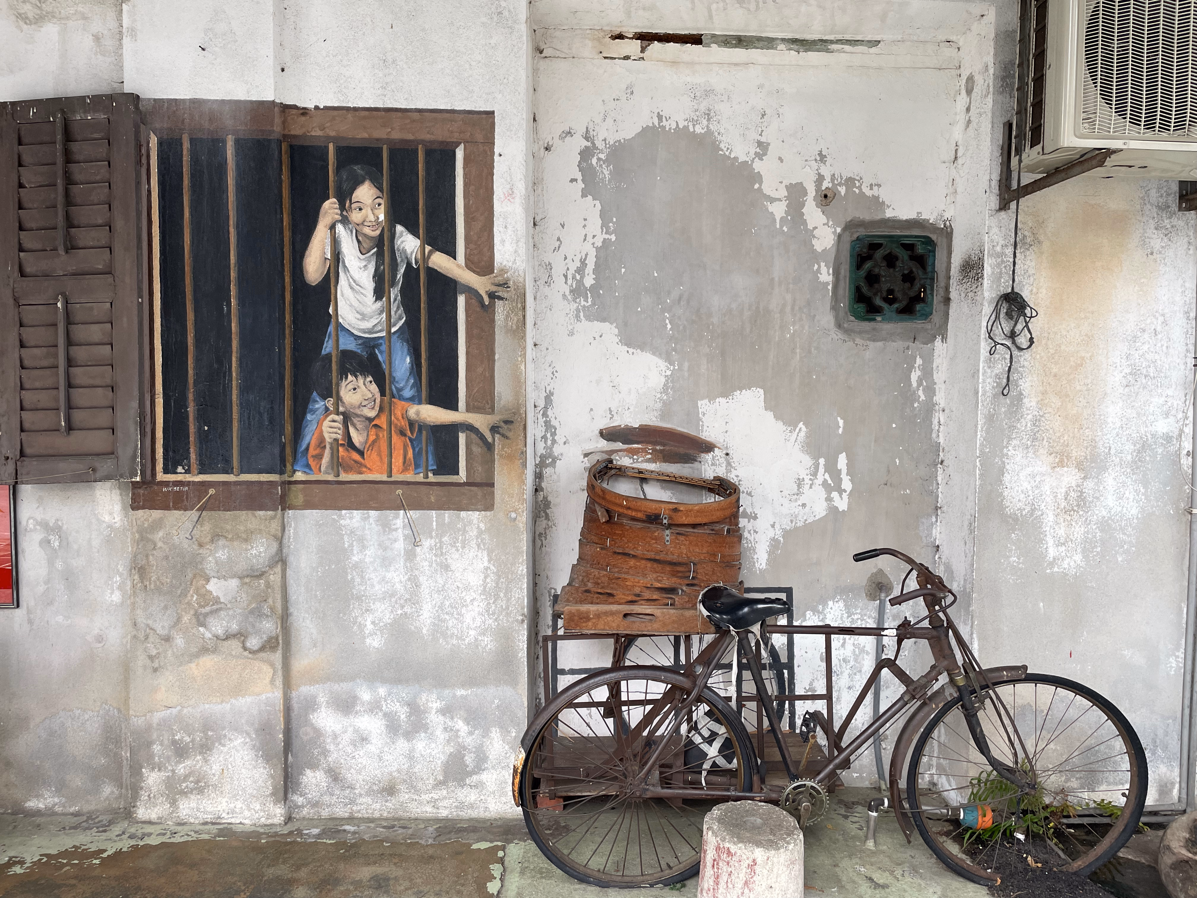 Two painted children lean through barred shutters, reaching toward a real rusted bicycle stacked with steamer baskets below.