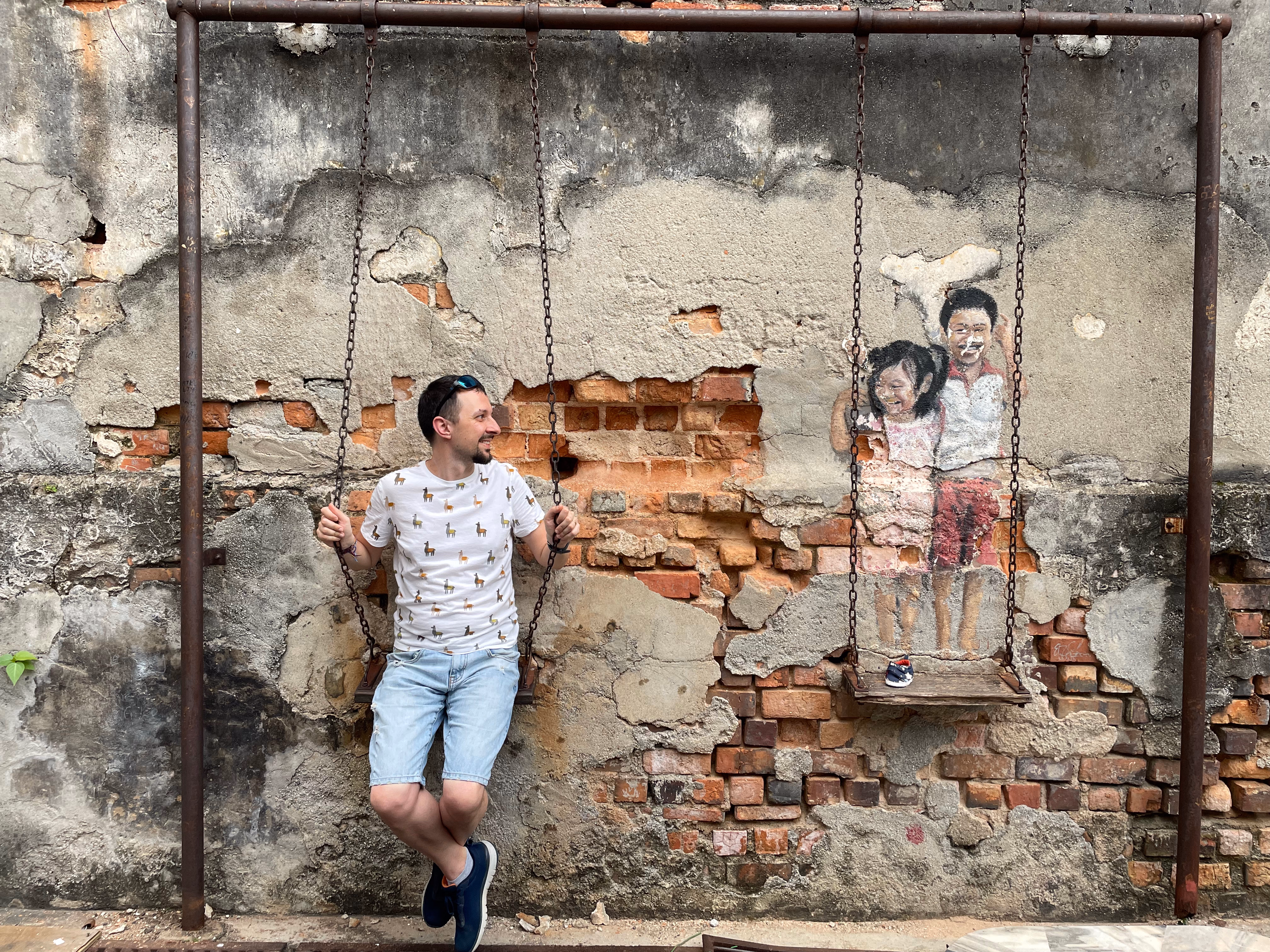 Alex on a metal swing installation next to a mural in George Town.