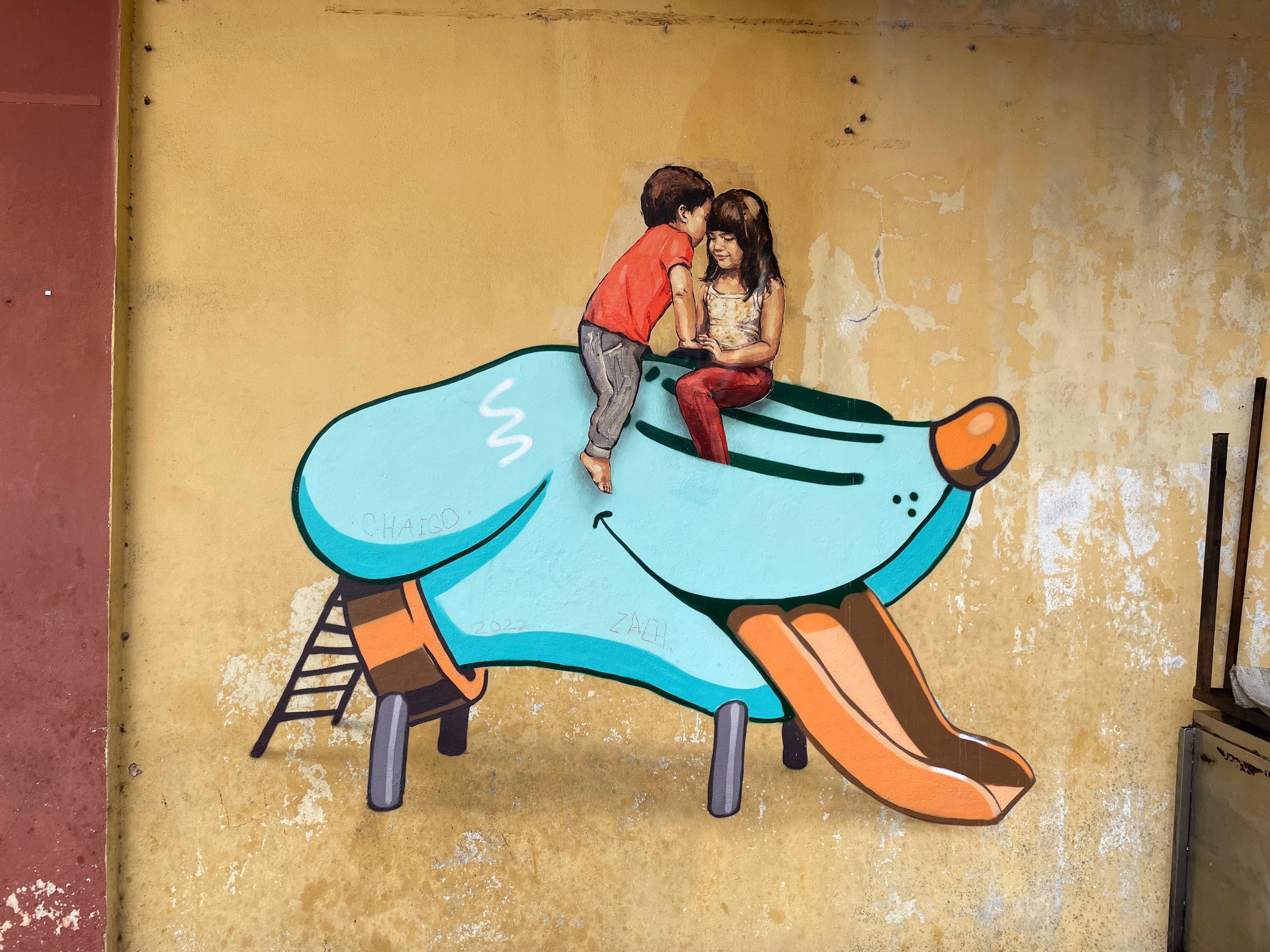 A boy whispers into a girl's ear as they sit atop a bright blue dog-shaped slide, its orange tongue curling down the chute.