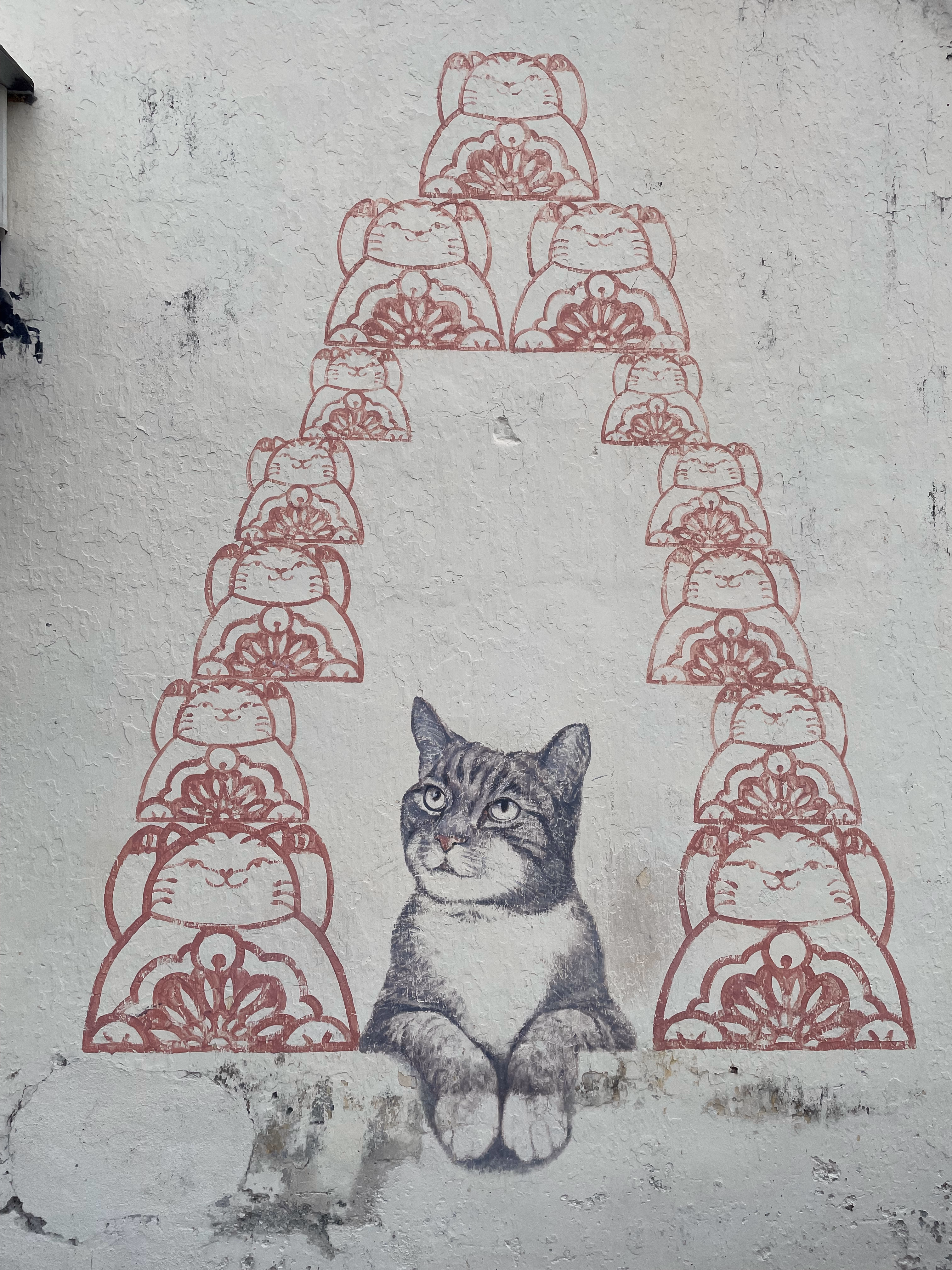 A tabby cat painted in greyscale looks up from a ledge, framed by a triangle of terracotta lucky-cat stamps on weathered stucco.