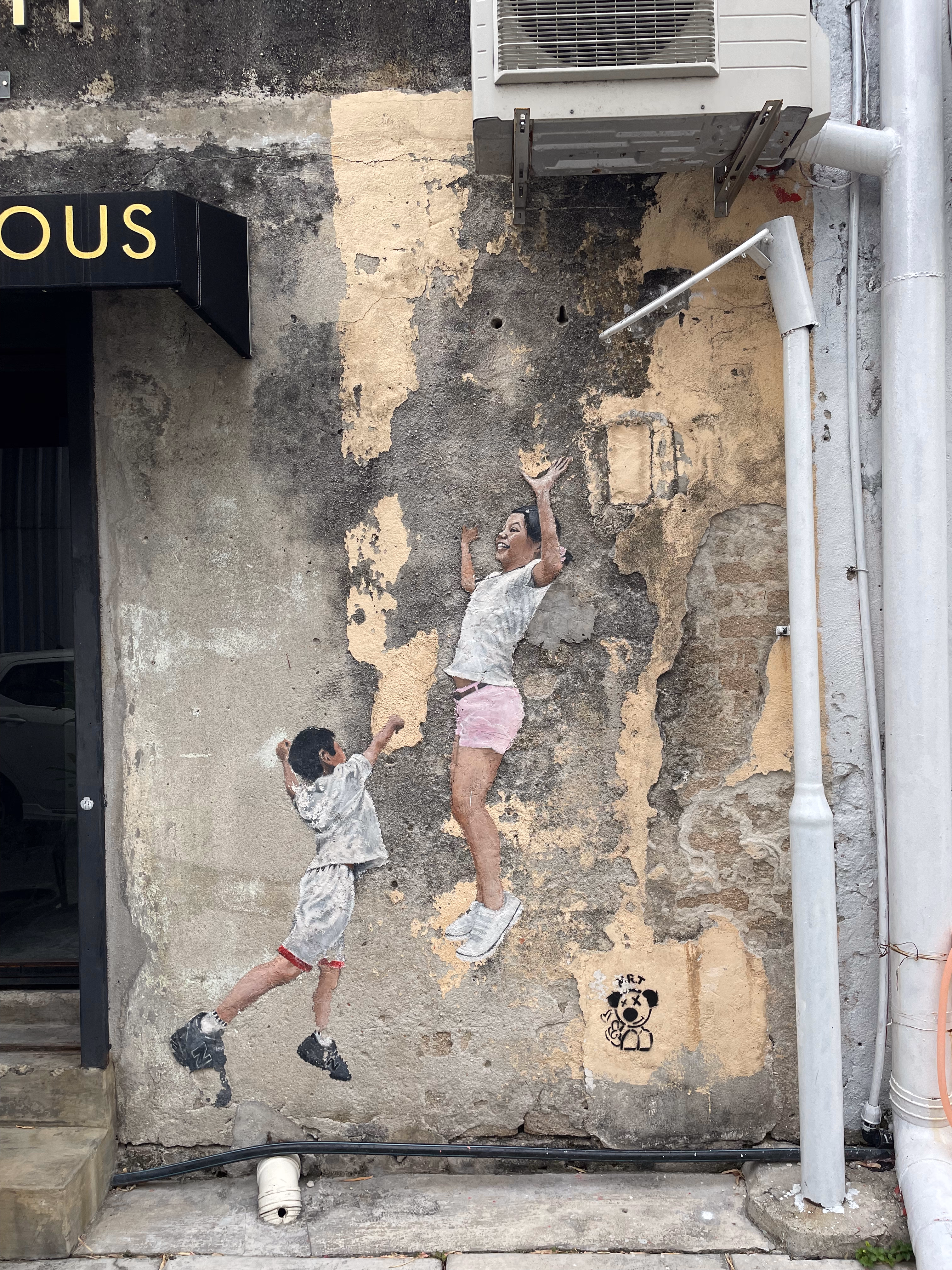 Two children painted mid-leap on a crumbling yellow wall, arms stretched upward against patches of exposed concrete.