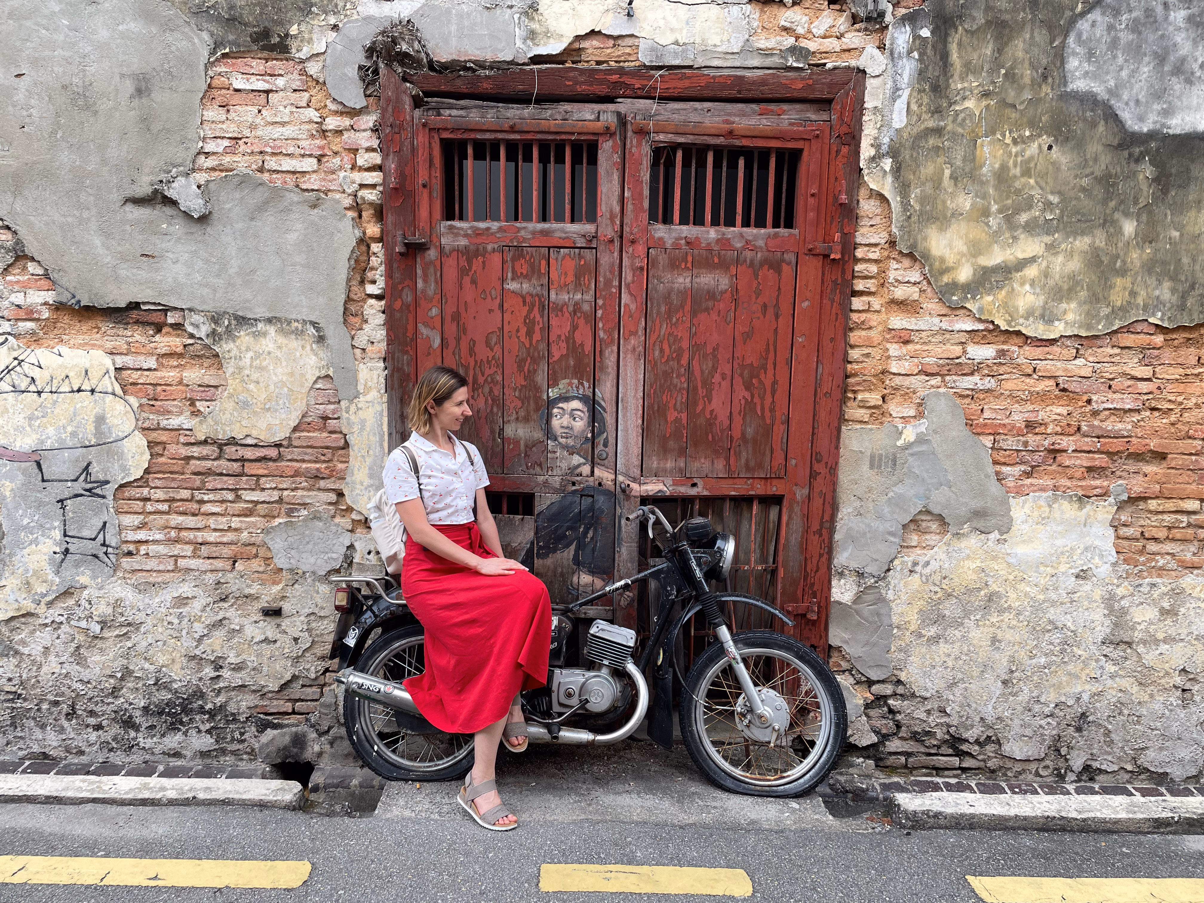 A real vintage motorcycle parked before red wooden doors, a painted boy in a helmet seated on the tank as if ready to ride off.