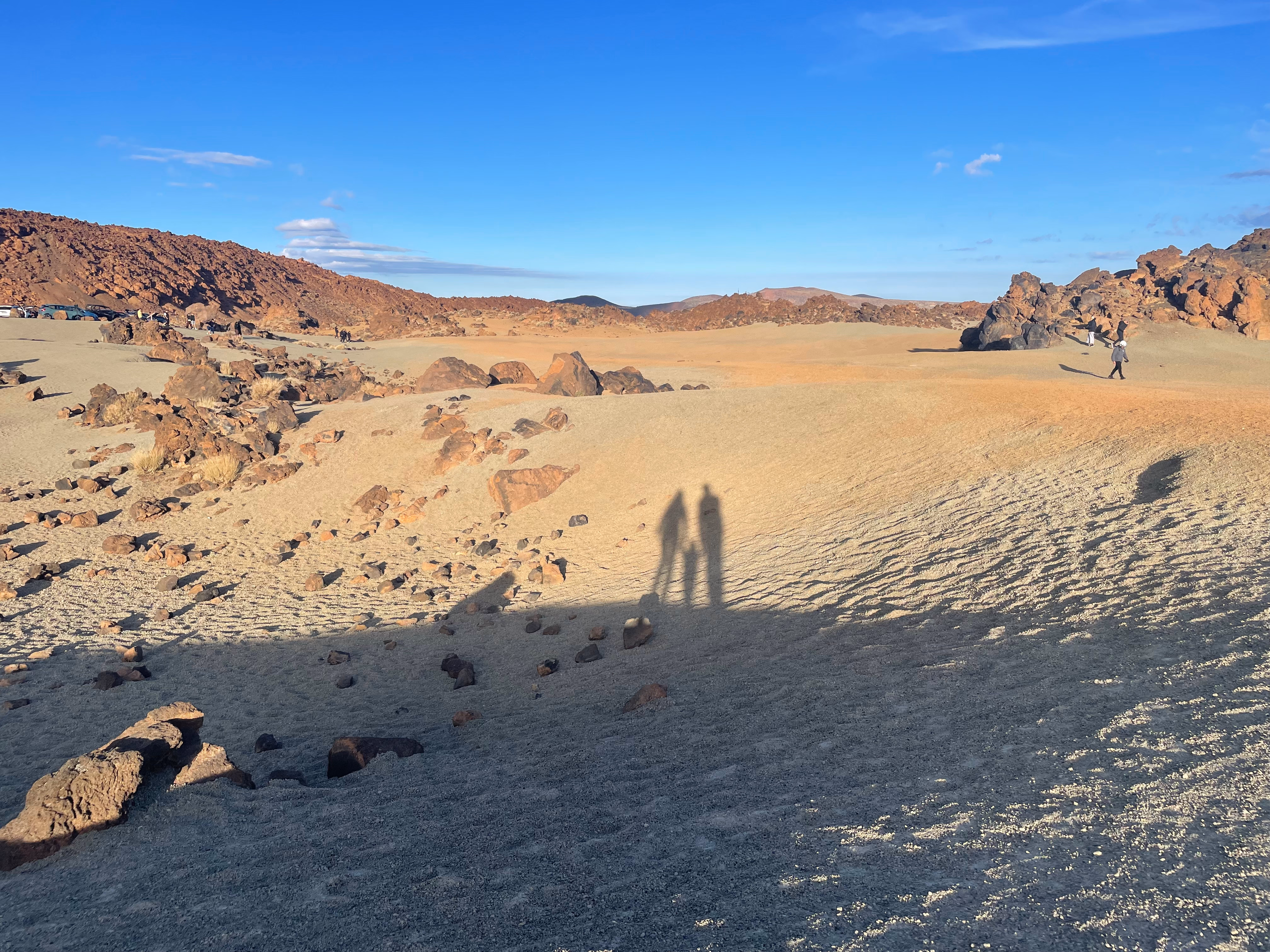 Late-afternoon light across the caldera floor — our shadows cast long on the ash, hikers and parked cars tiny against the dark volcanic hills beyond.