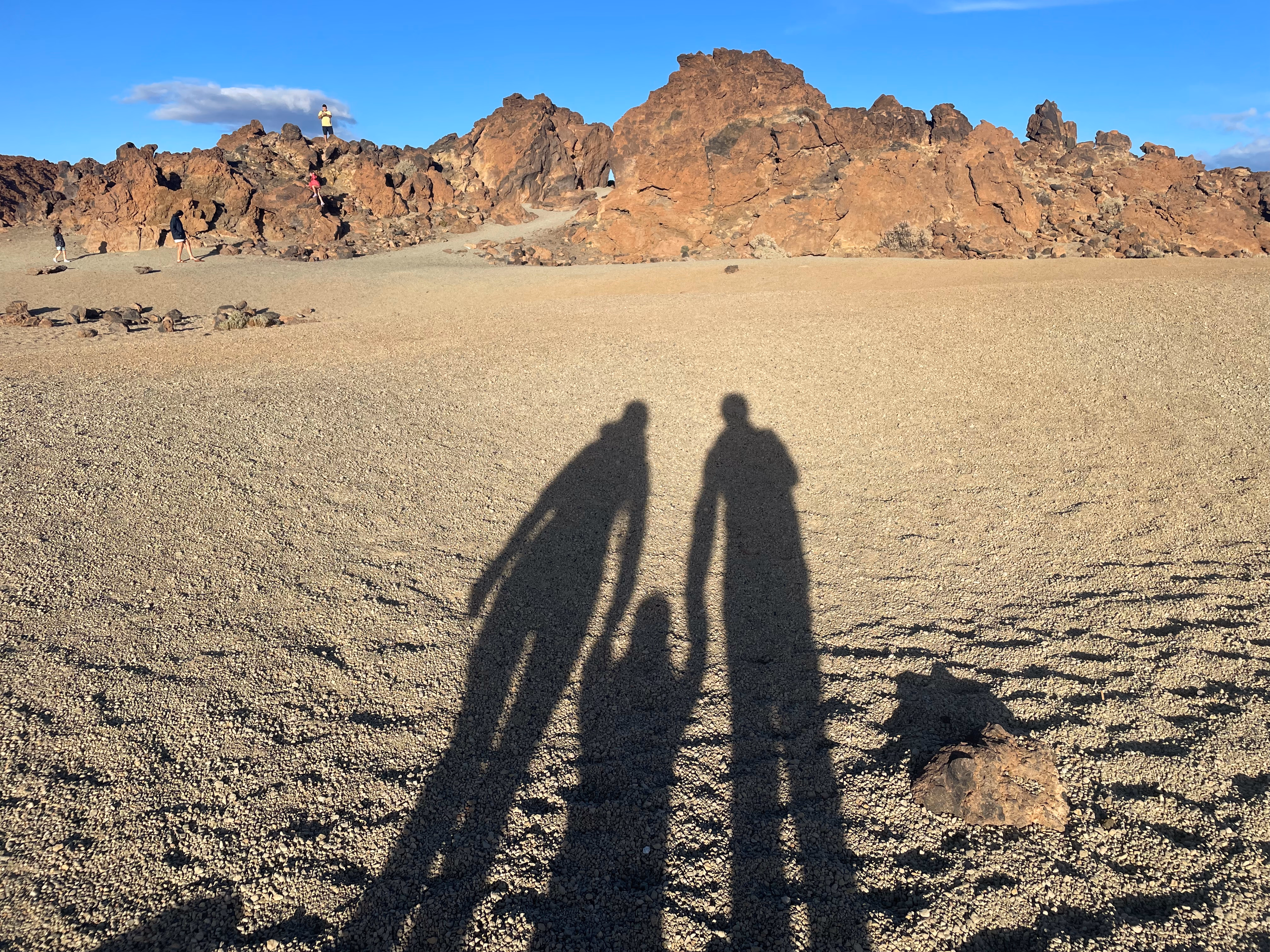 Three shadows — two adults and a child between them — stretch across light volcanic gravel toward a jagged reddish ridge where other walkers pick their way along the rocks.