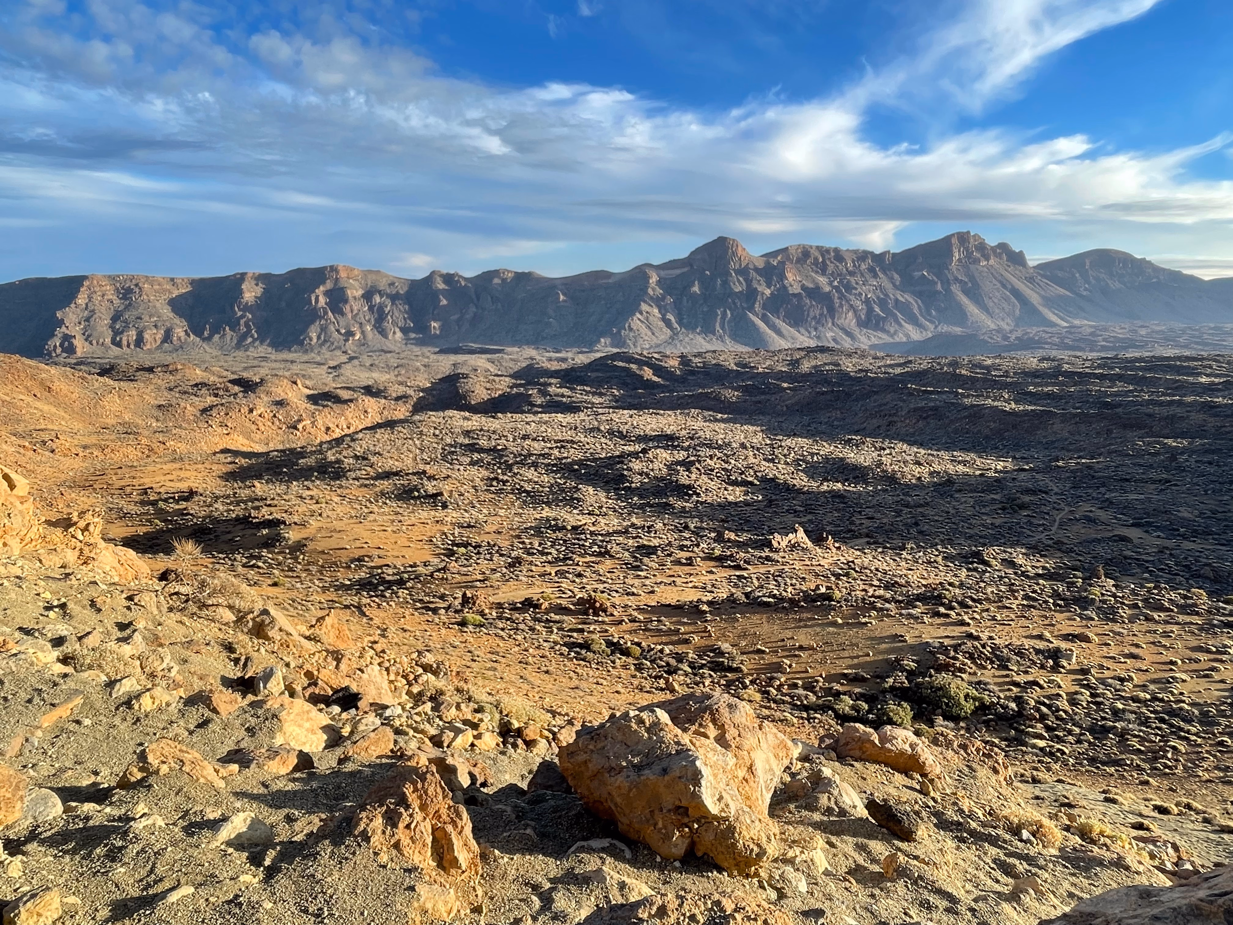 Wide open caldera under wispy cloud — pale ash and scattered boulders in the foreground, a dark rocky ridge stretching across the horizon in late-afternoon light.