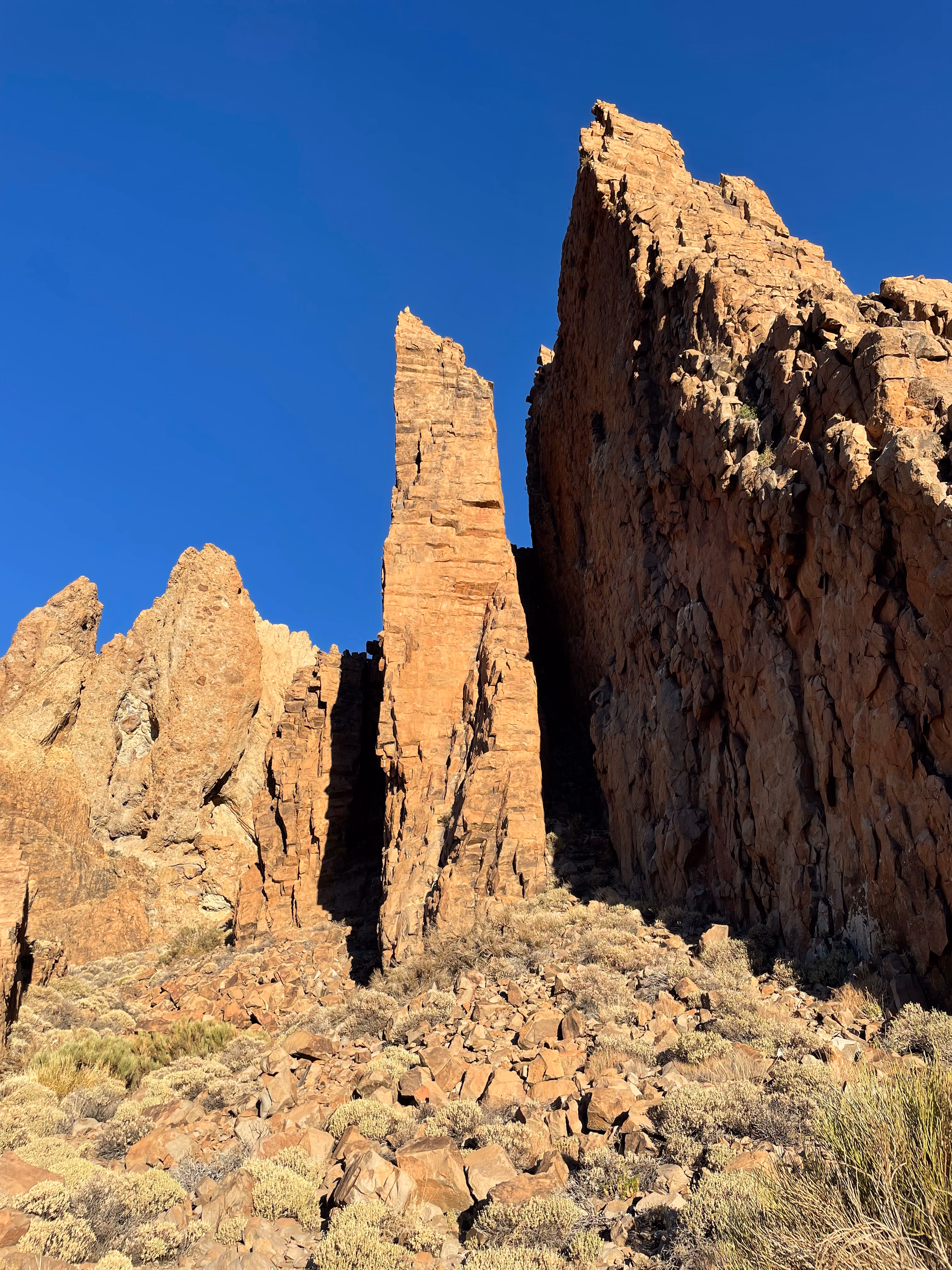 Needle-thin volcanic spires catching golden light — warm orange faces against deep blue sky, loose rubble and desert scrub at their base.