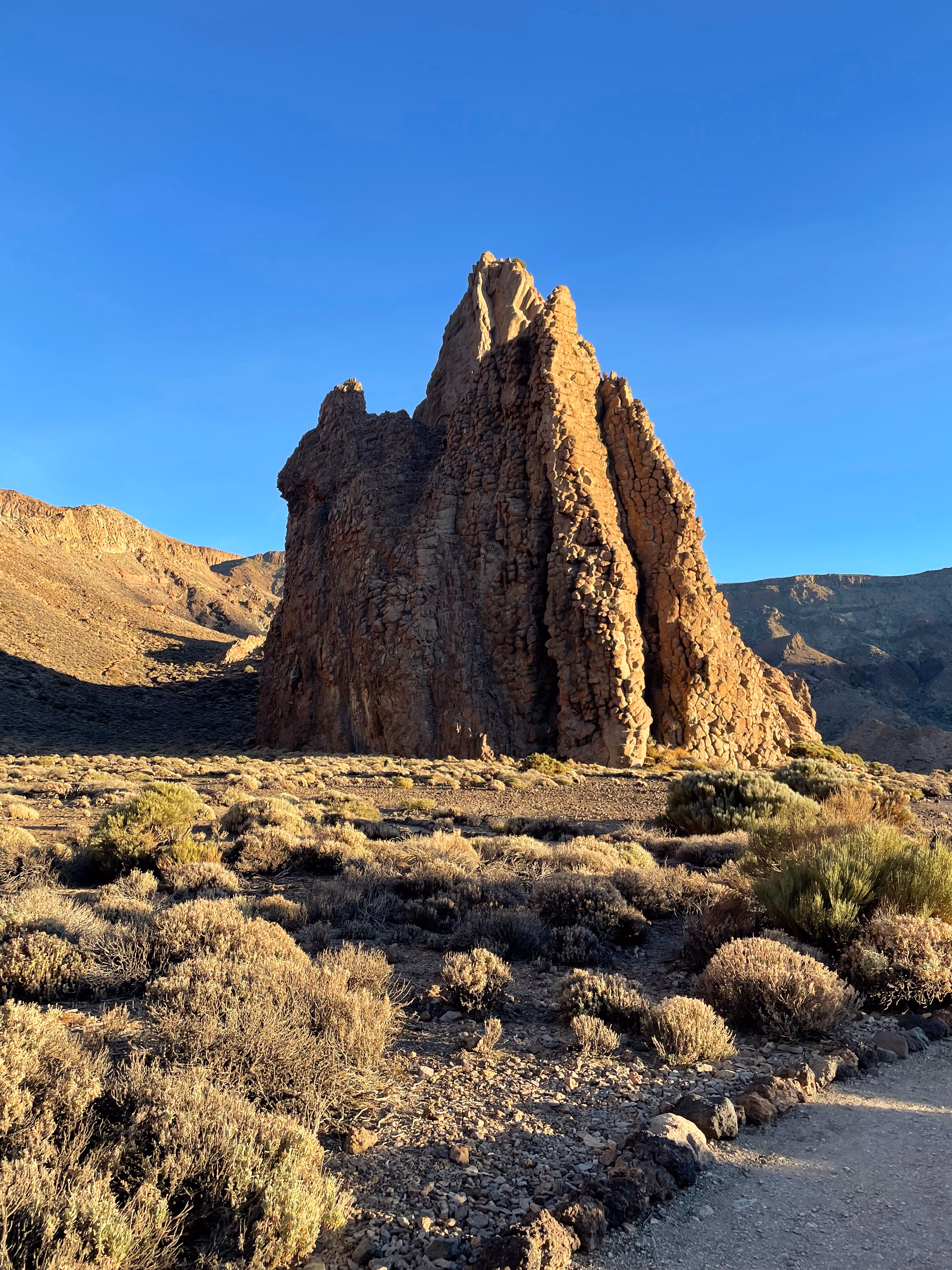 A solitary reddish pillar rises from a plain of rounded shrubs — one face lit, the other in shadow, a gravel path edged with dark lava rocks in the corner.
