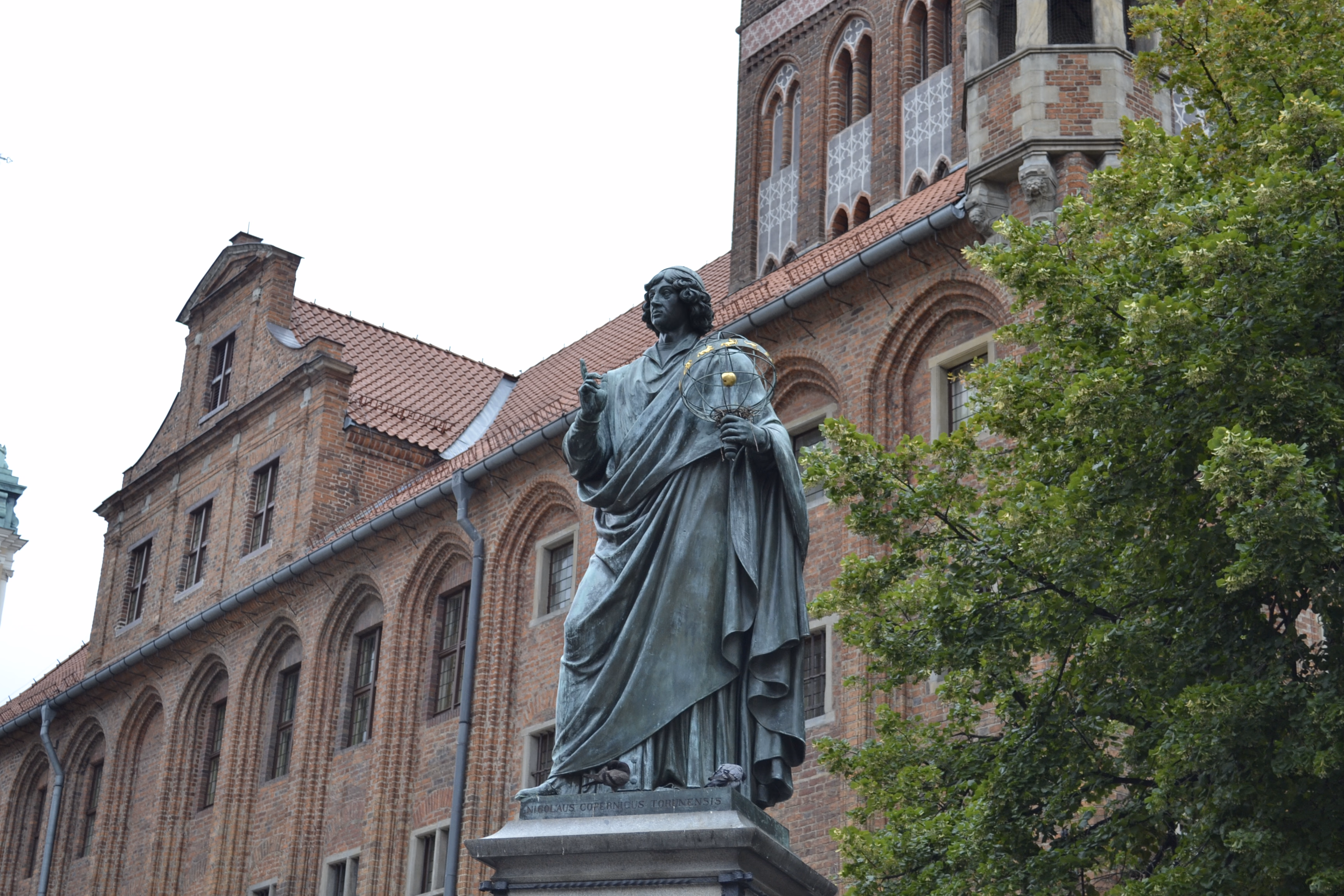 Statue of Copernicus in Toruń — a Brick Gothic cathedral dome above a carpet of red-tiled roofs and cream plastered walls.