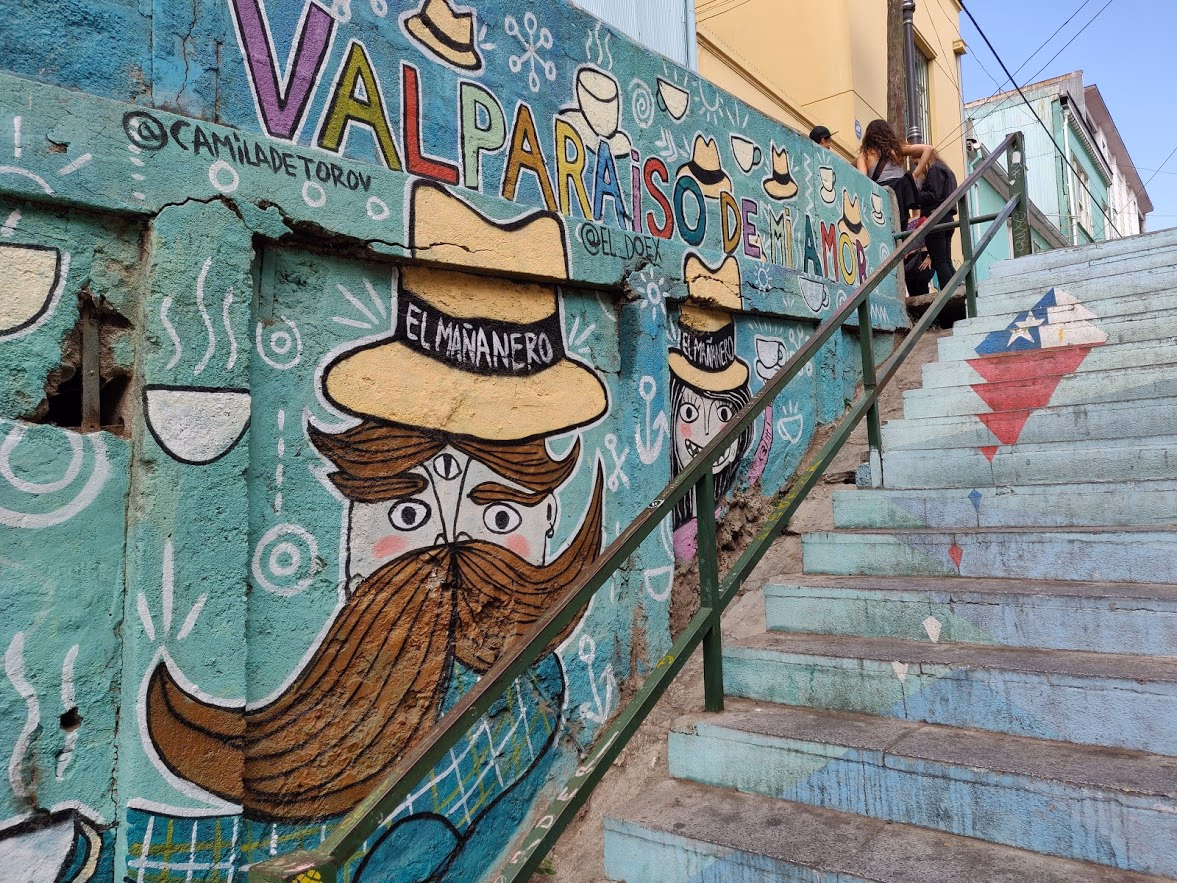 Concrete stairs painted in blue and white lead past a mural spelling "Valparaíso de mi amor" — a mustachioed figure in a yellow hat above a Chilean flag on the risers.