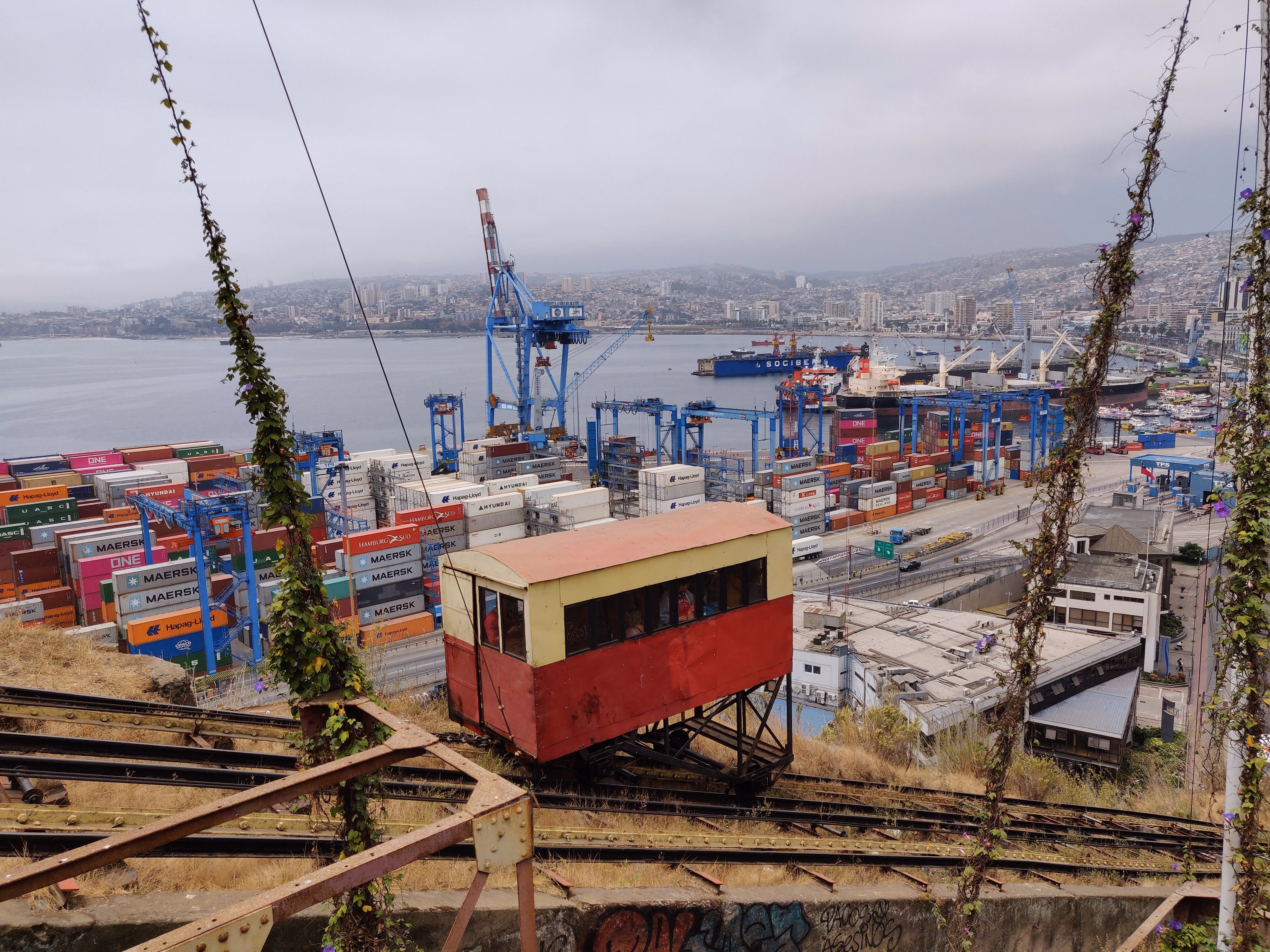 A cream-and-red funicular car clings to its track above the port — stacks of Maersk, ONE and Hamburg Süd containers and gantry cranes stretching to the bay.